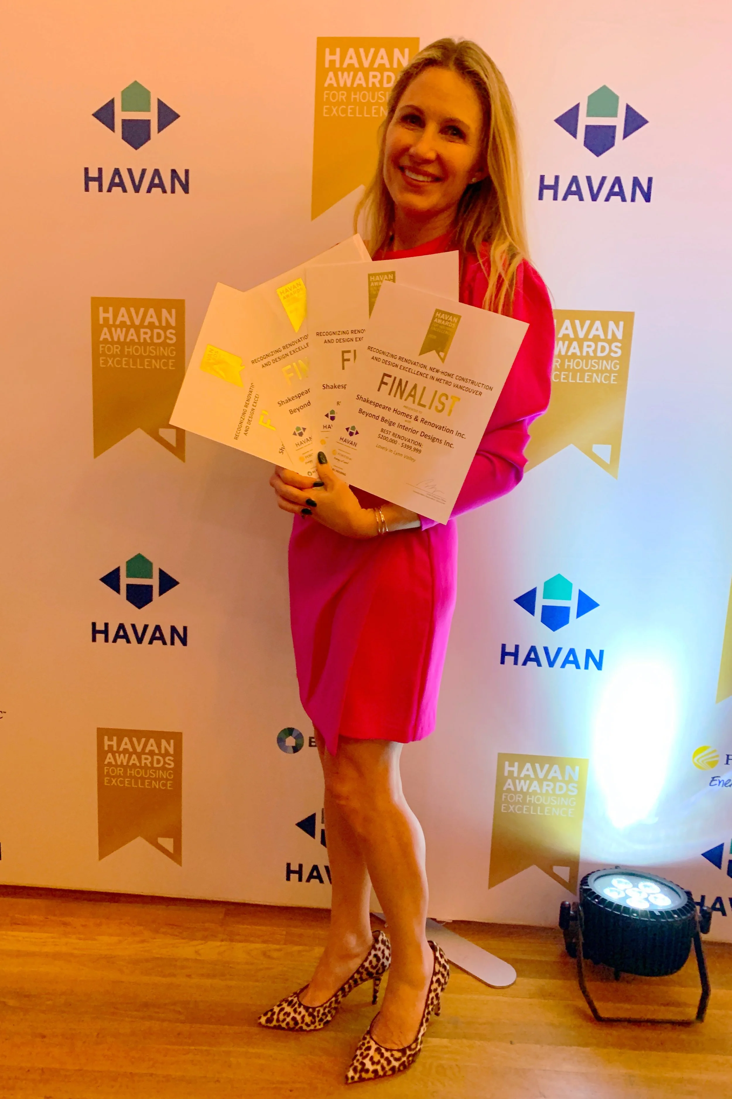 A woman in a bright pink dress holding four award certificates, standing on a wooden floor in front of a backdrop that displays multiple 'Havan Awards for Housing Excellence' logos. She is wearing leopard print high heels and smiling.
