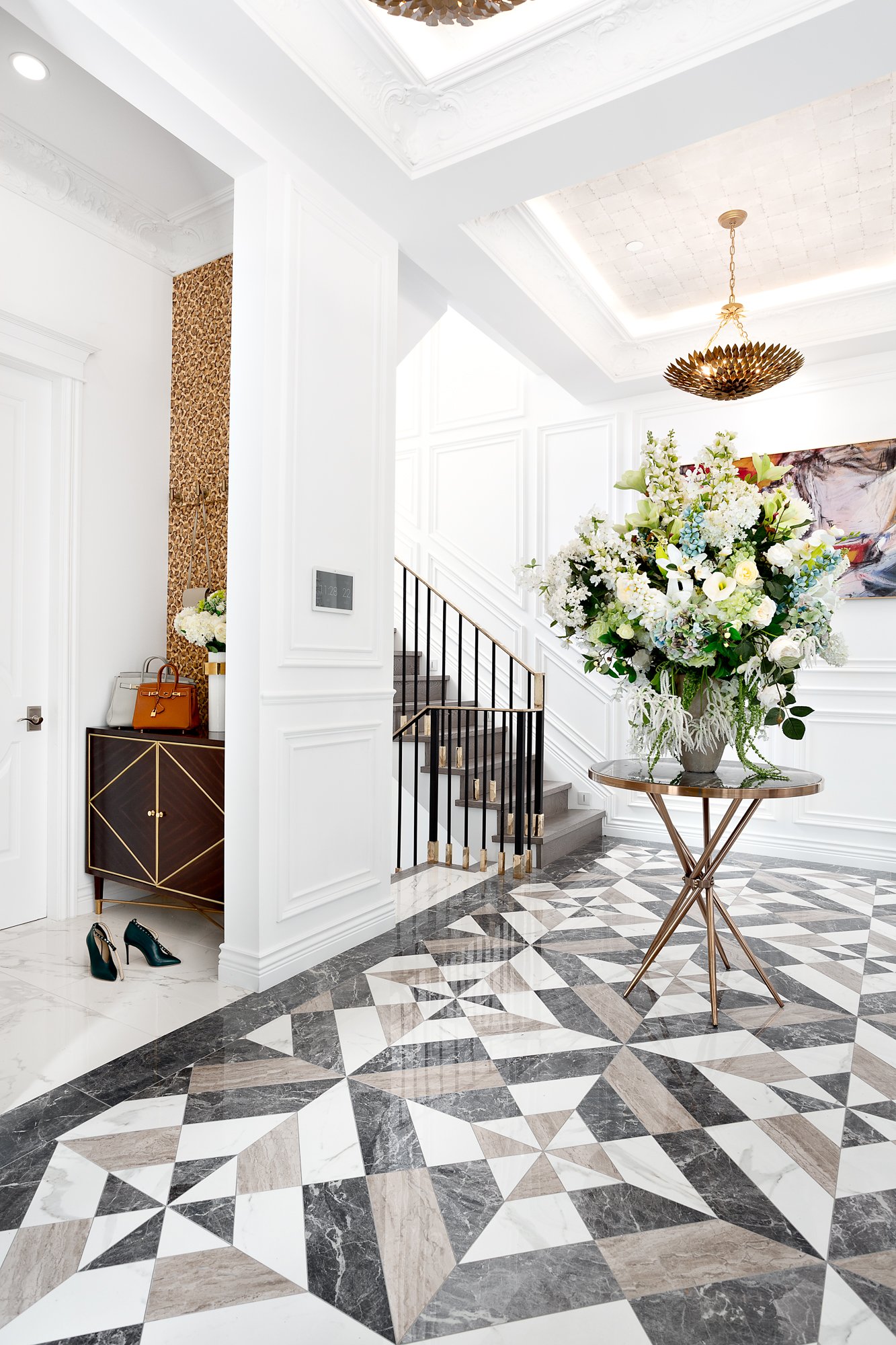 Grand foyer in a Vancouver luxury home featuring a bold geometric tiled floor, crisp white paneled walls, and a gold pedestal table with a large floral arrangement.