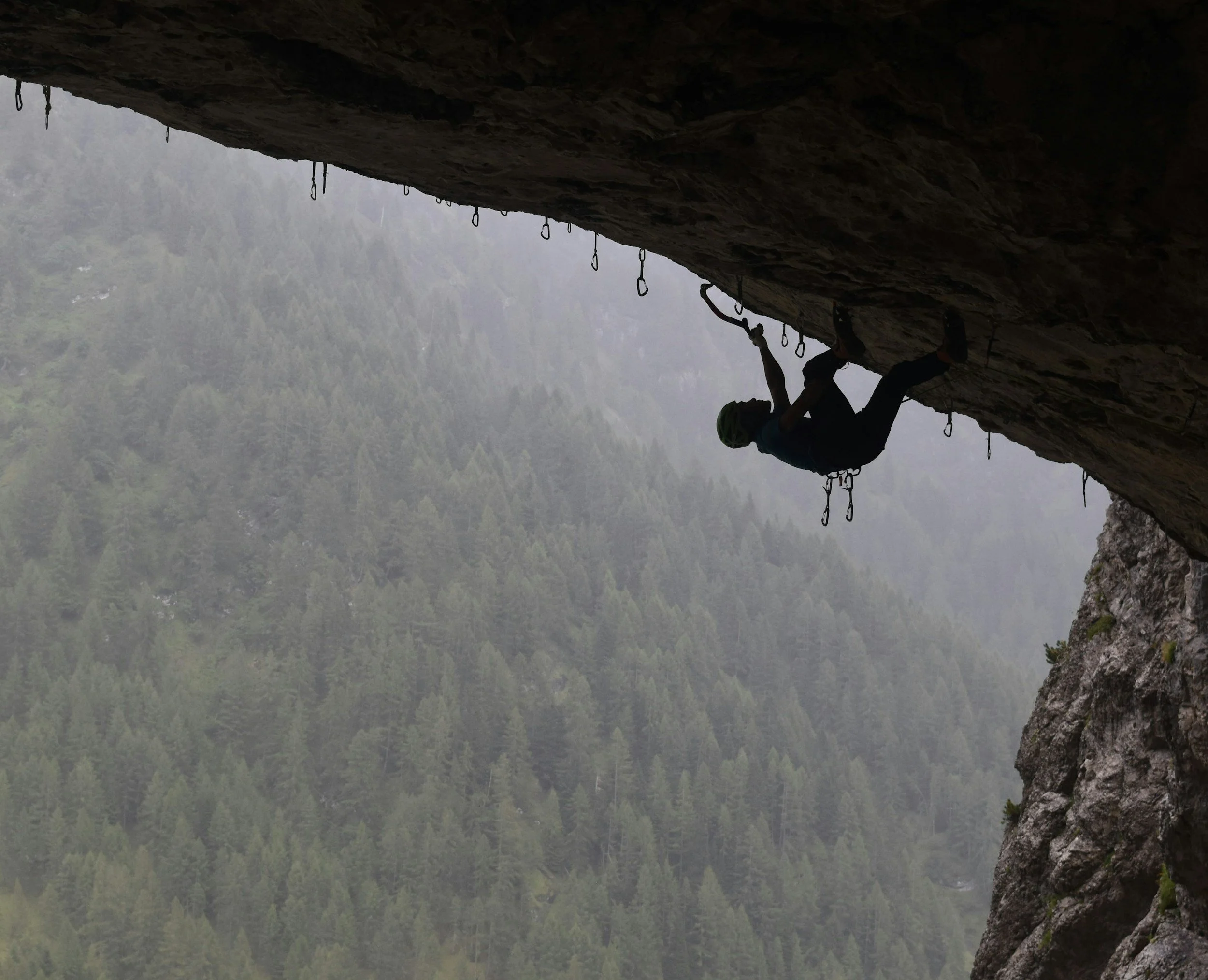 A man climbing a nearly horizontal section of a cliff, high above a forest.