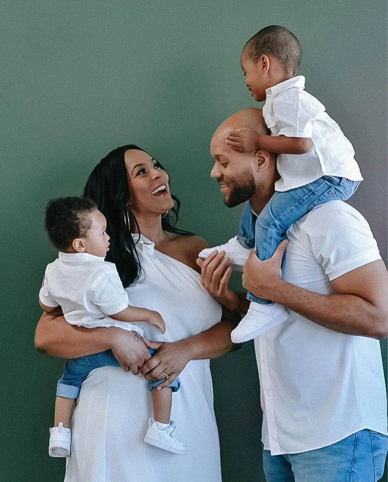 A joyful family of four with a woman, man, and two young boys, all smiling and interacting affectionately against a green background.
