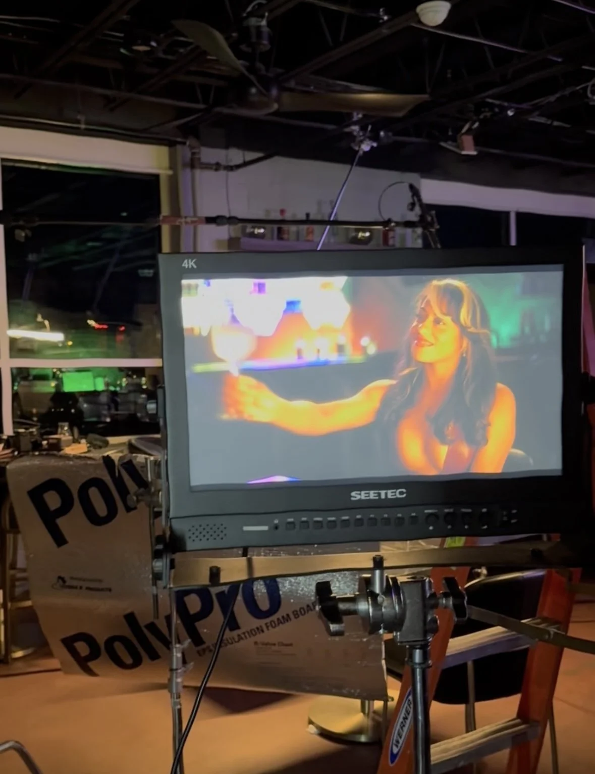 Coley holding a drink in a bar setting, illuminated by colorful neon lighting, on a filmmaking or video production set.