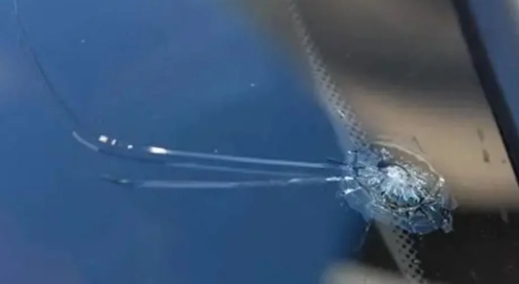 Close-up of a cracked windshield with a bullet hole, showing spiderweb cracks emanating from the impact point.