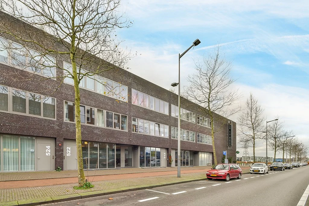 Street view of a modern four-story brick building with large windows, leafless trees along the sidewalk, parked cars, and a blue sky with scattered clouds.