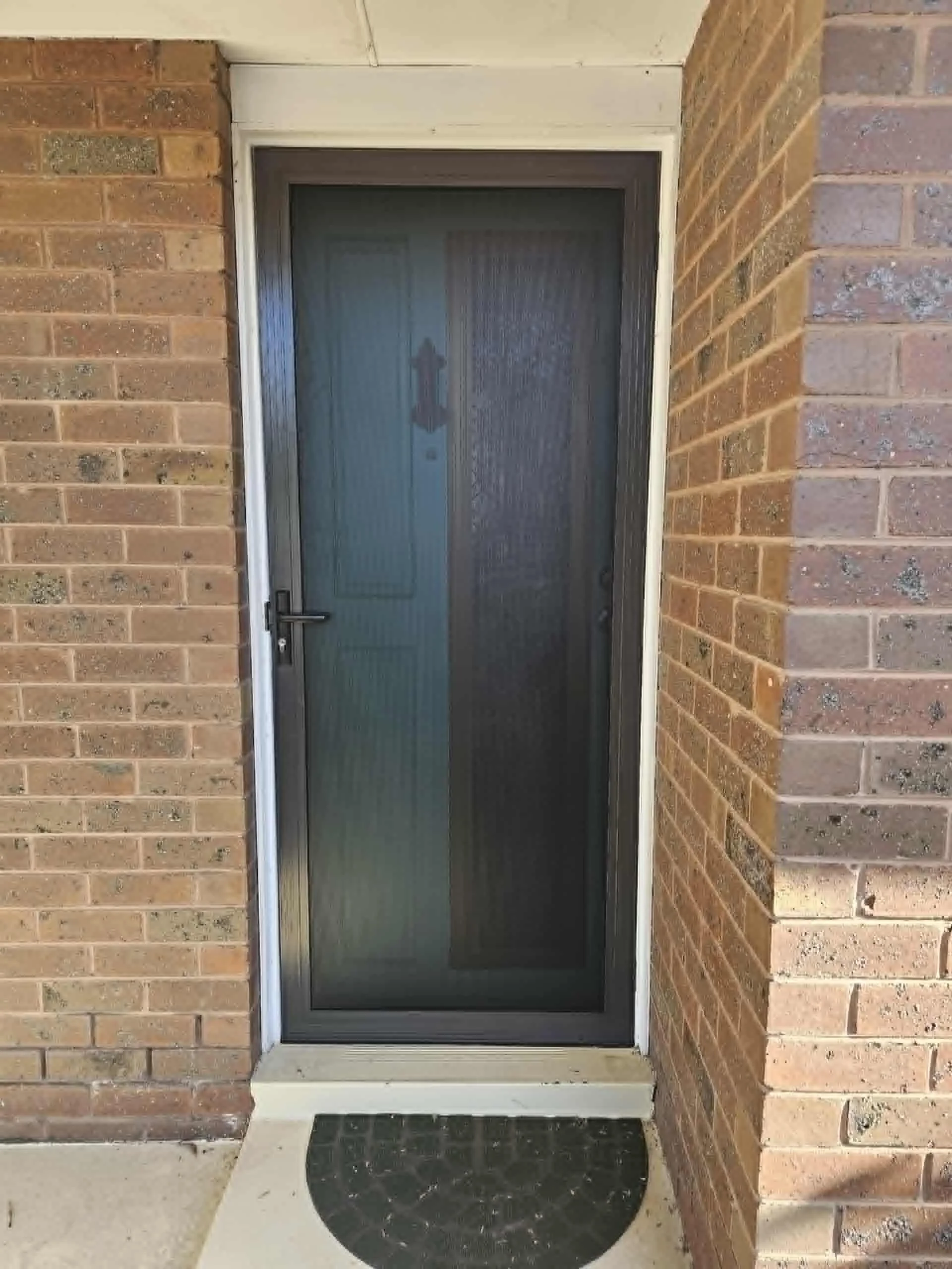 Front door with a black security screen door and a brick wall on the sides, with a small black doormat in front.