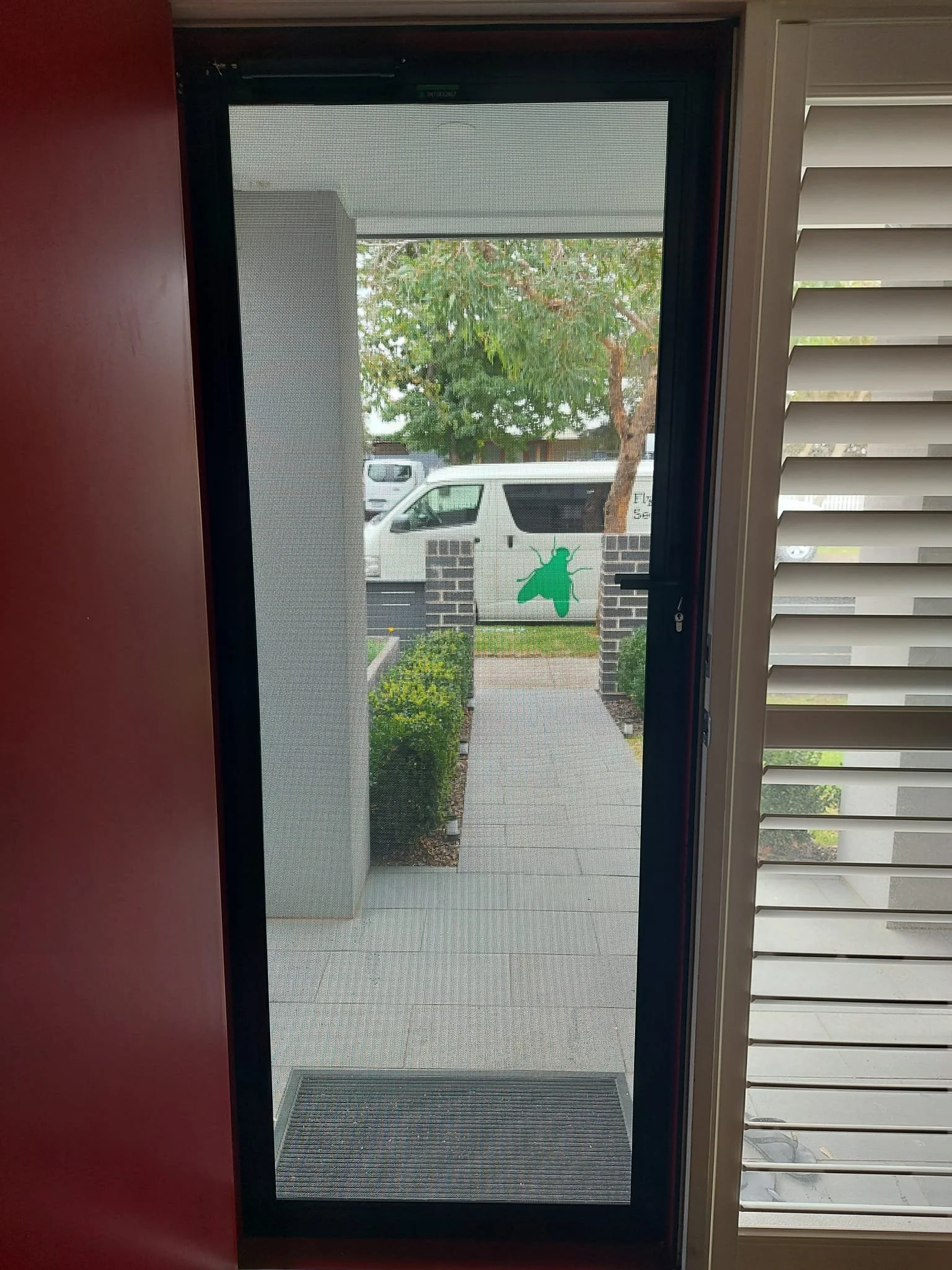 View through a glass door showing a sidewalk, a brick wall with a green insect decal, and parked vehicles including a white van with a green insect logo