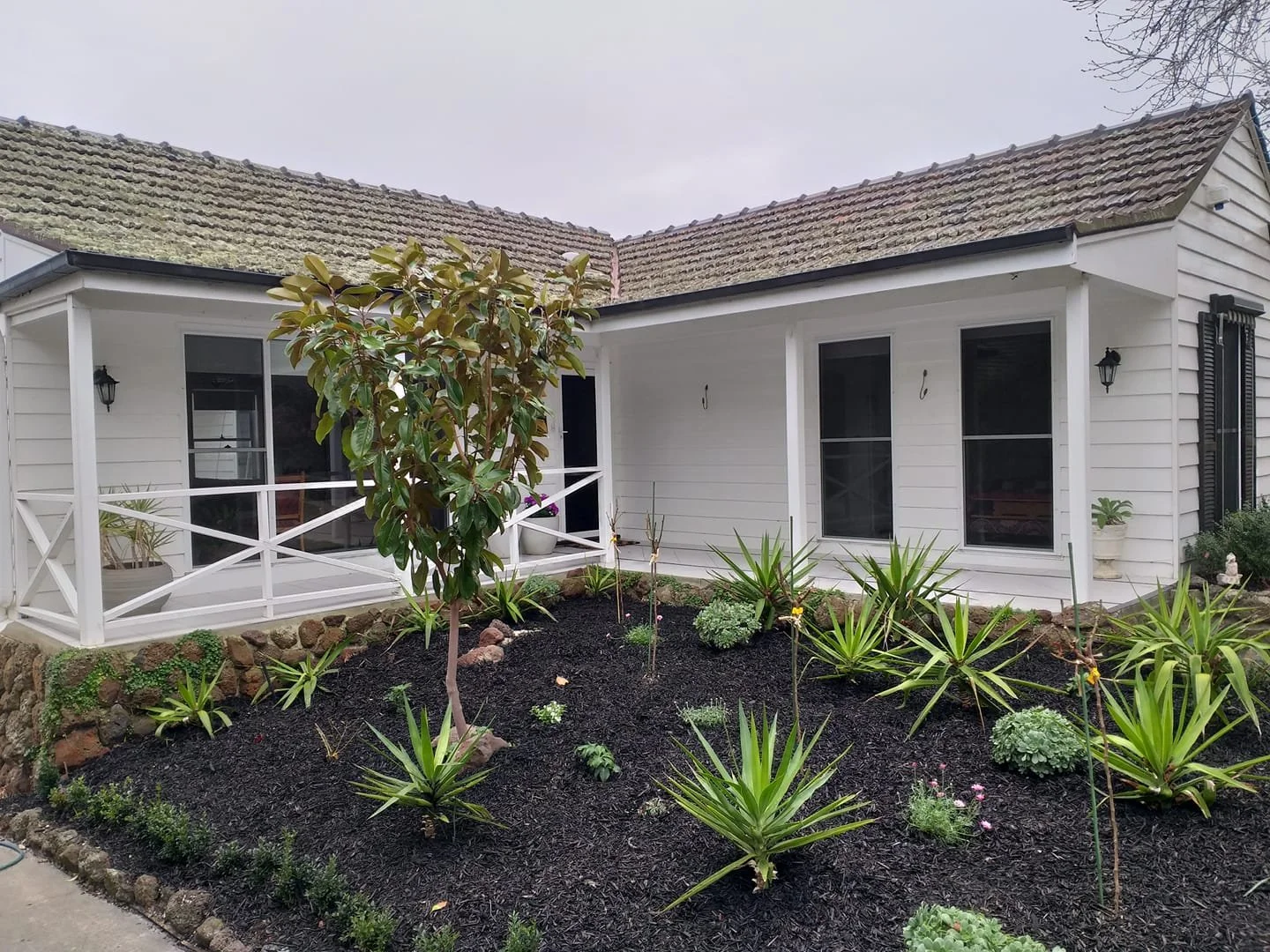White house with a front porch, black shutters, and a garden with green plants and mulch in the front yard.