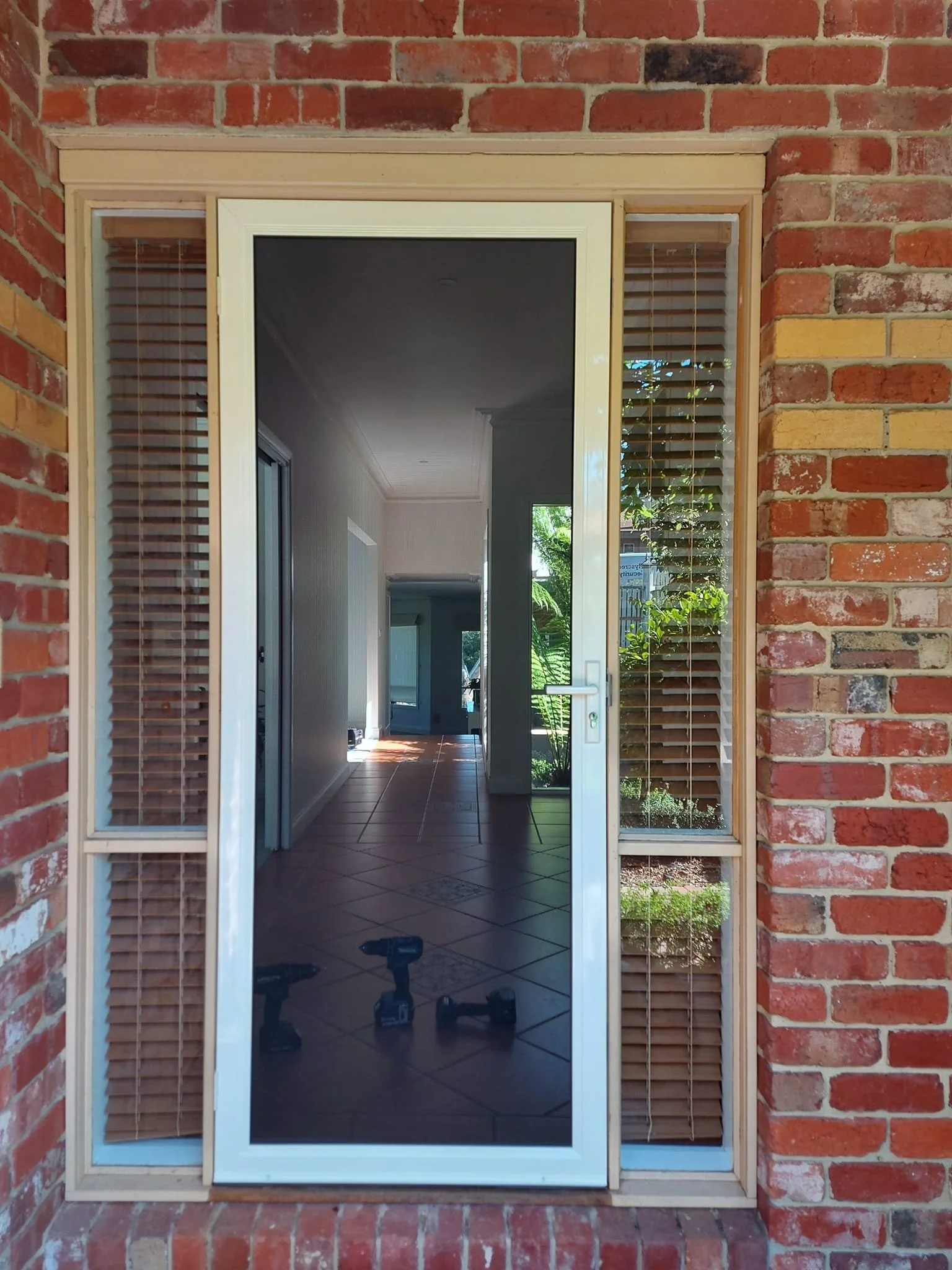 Front door with glass panel and wooden blinds, brick exterior wall, view inside a hallway with tiled flooring, tools on the floor, and lush greenery outside