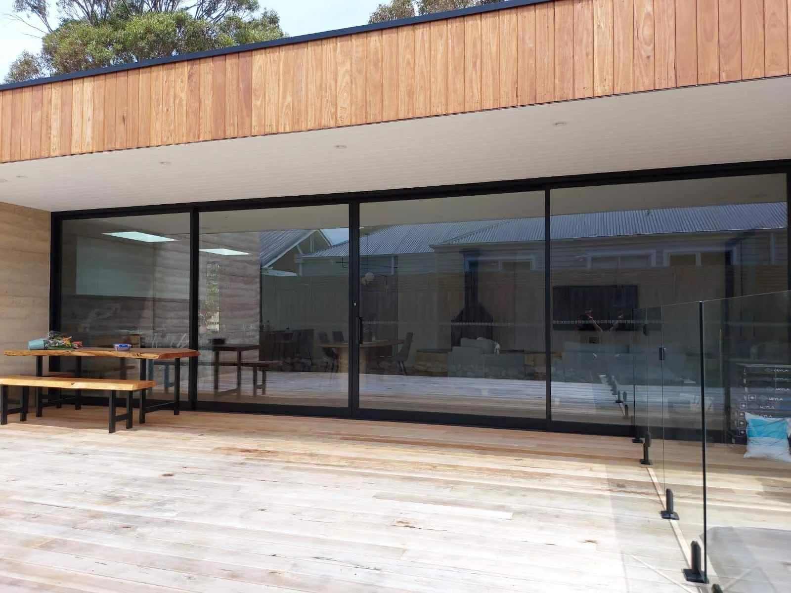 Modern house with large glass sliding doors on wooden deck, patio furniture, and a glass railing.