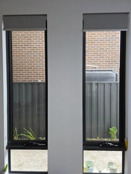 Two tall windows with blinds, showing a brick wall, a metal fence, and a small plant outside.