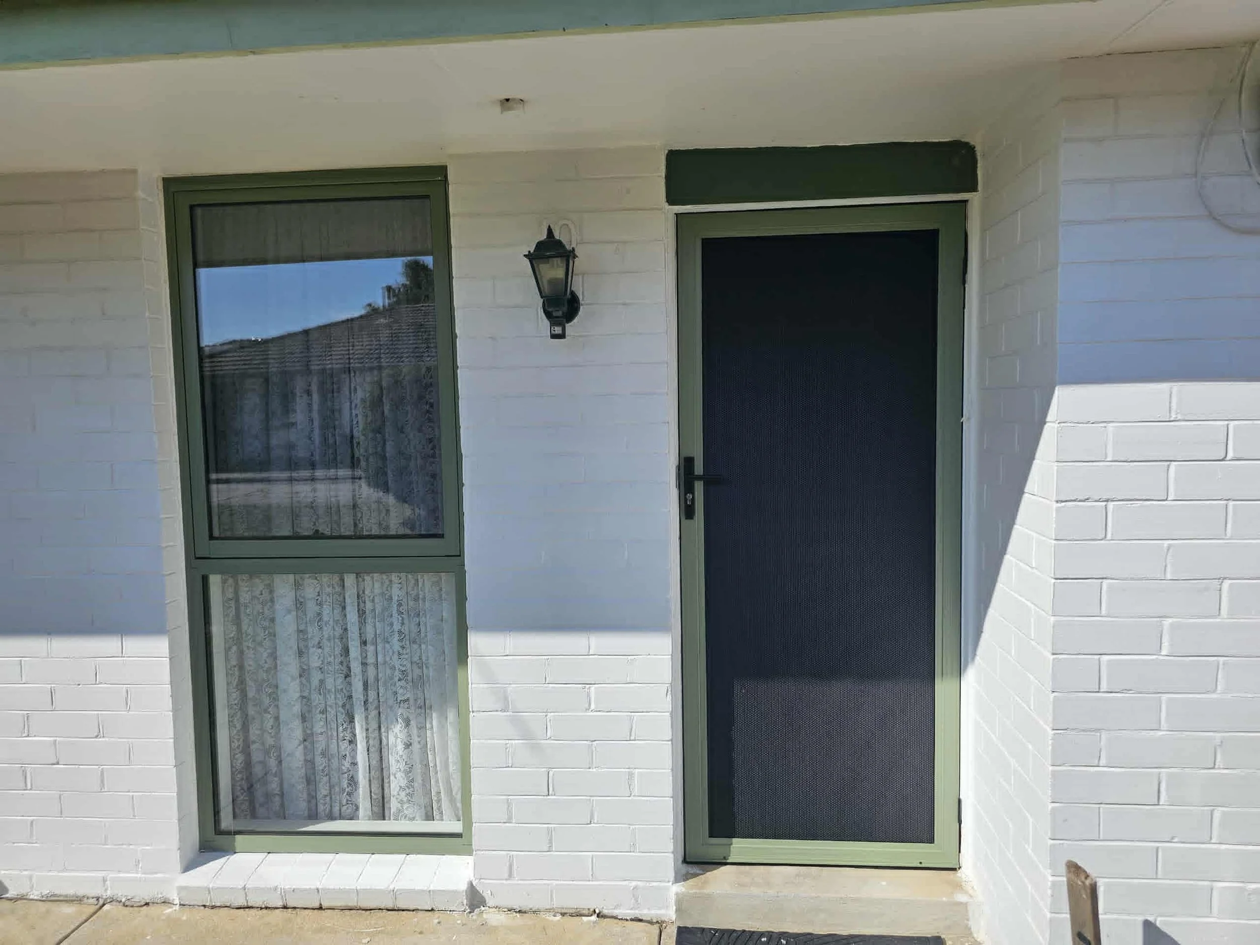 Front door and window of a white brick house with a green door and window frame, and an outdoor wall-mounted lantern light.