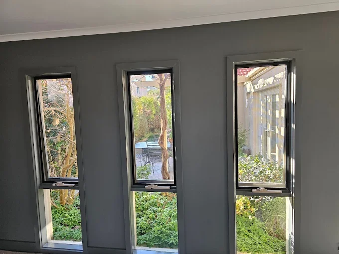 Three tall, narrow windows in a gray wall, showing a backyard with trees, bushes, and a patio table.
