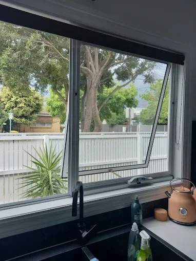 View from inside a kitchen showing a window with a view of trees, a white fence, and neighboring houses.