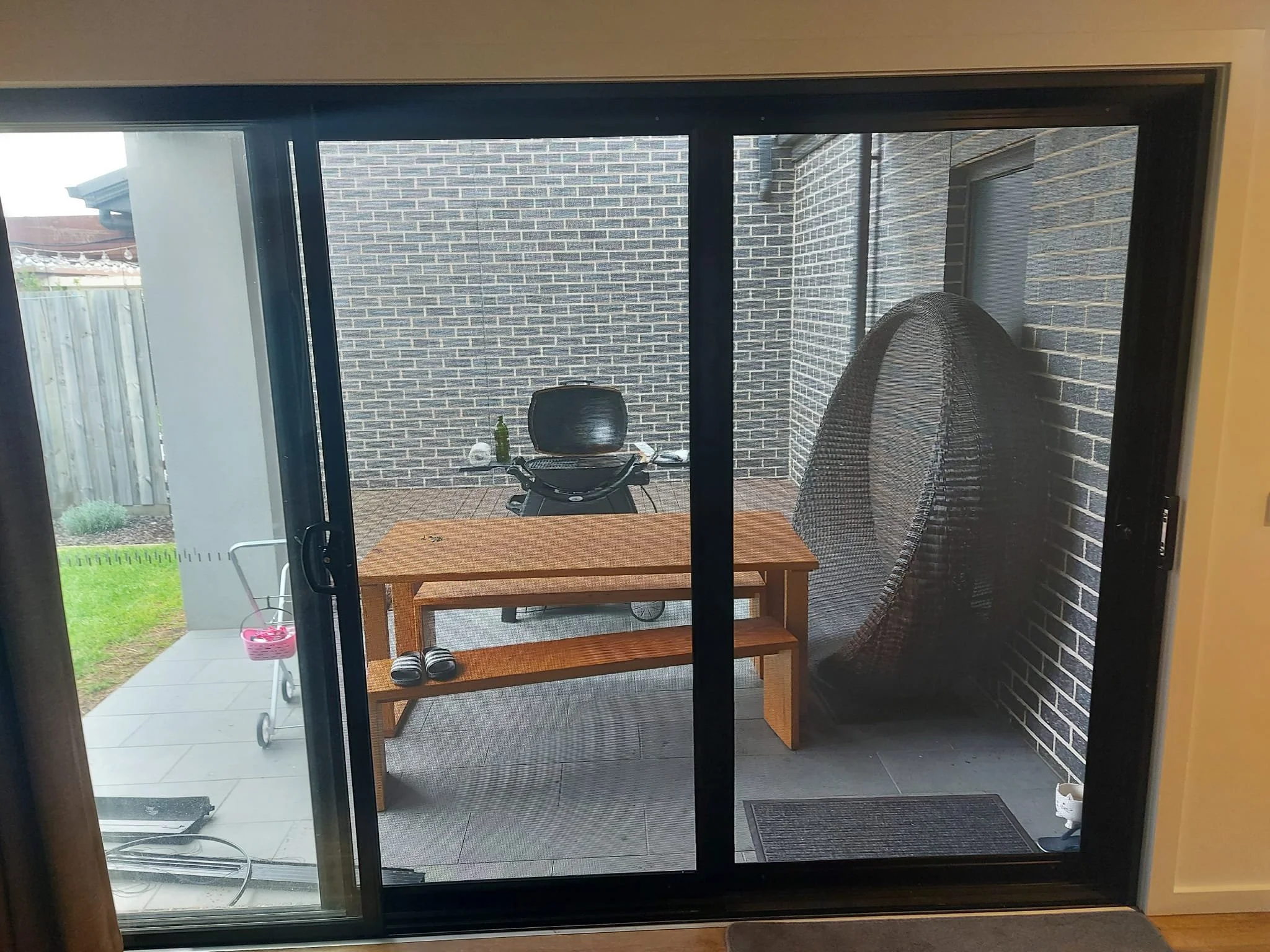 Outdoor patio area viewed through a sliding glass door, with a wooden table, a pair of slippers, a large black wicker egg-shaped chair, and a barbecue grill, surrounded by brick walls.