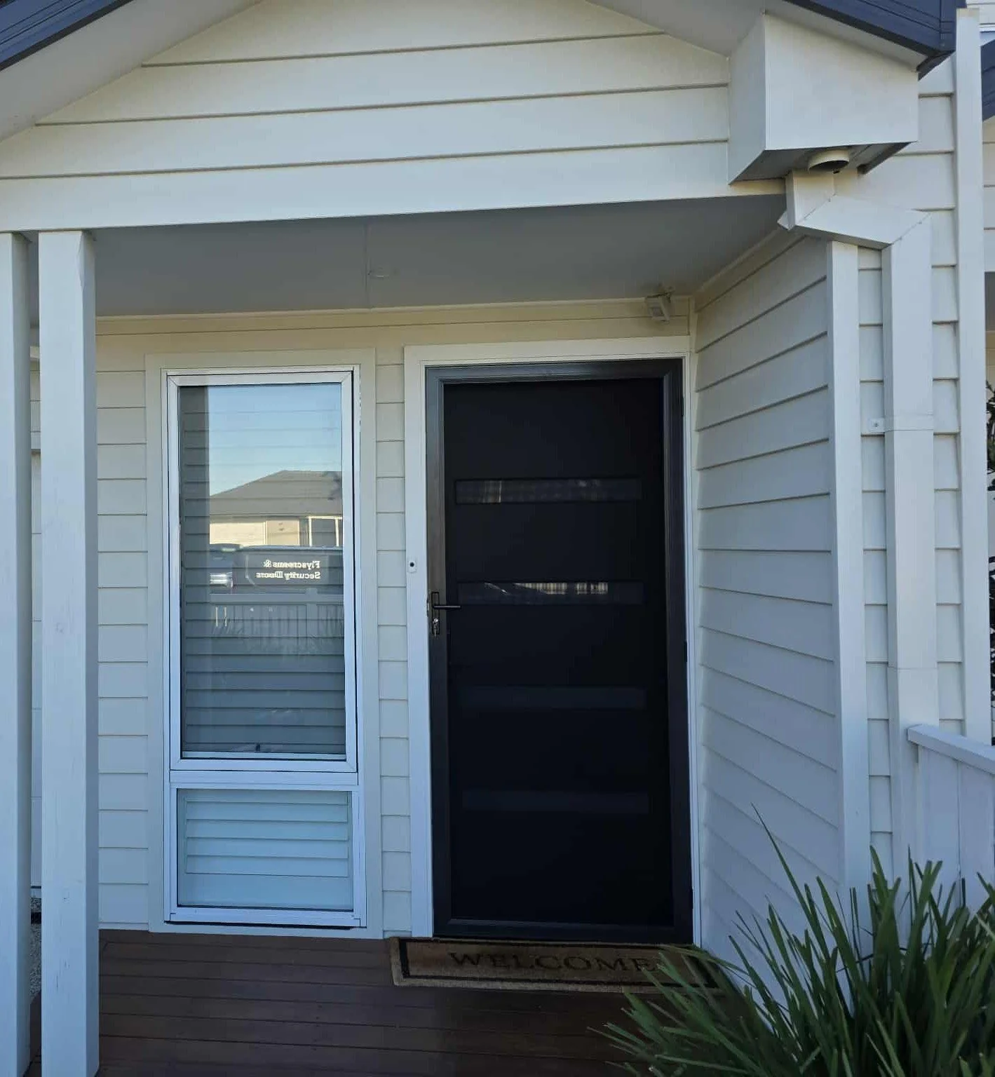 The front porch of a house with a white exterior, featuring a screened door and a glass window, with a welcome mat on the wooden floor and some plants at the side.