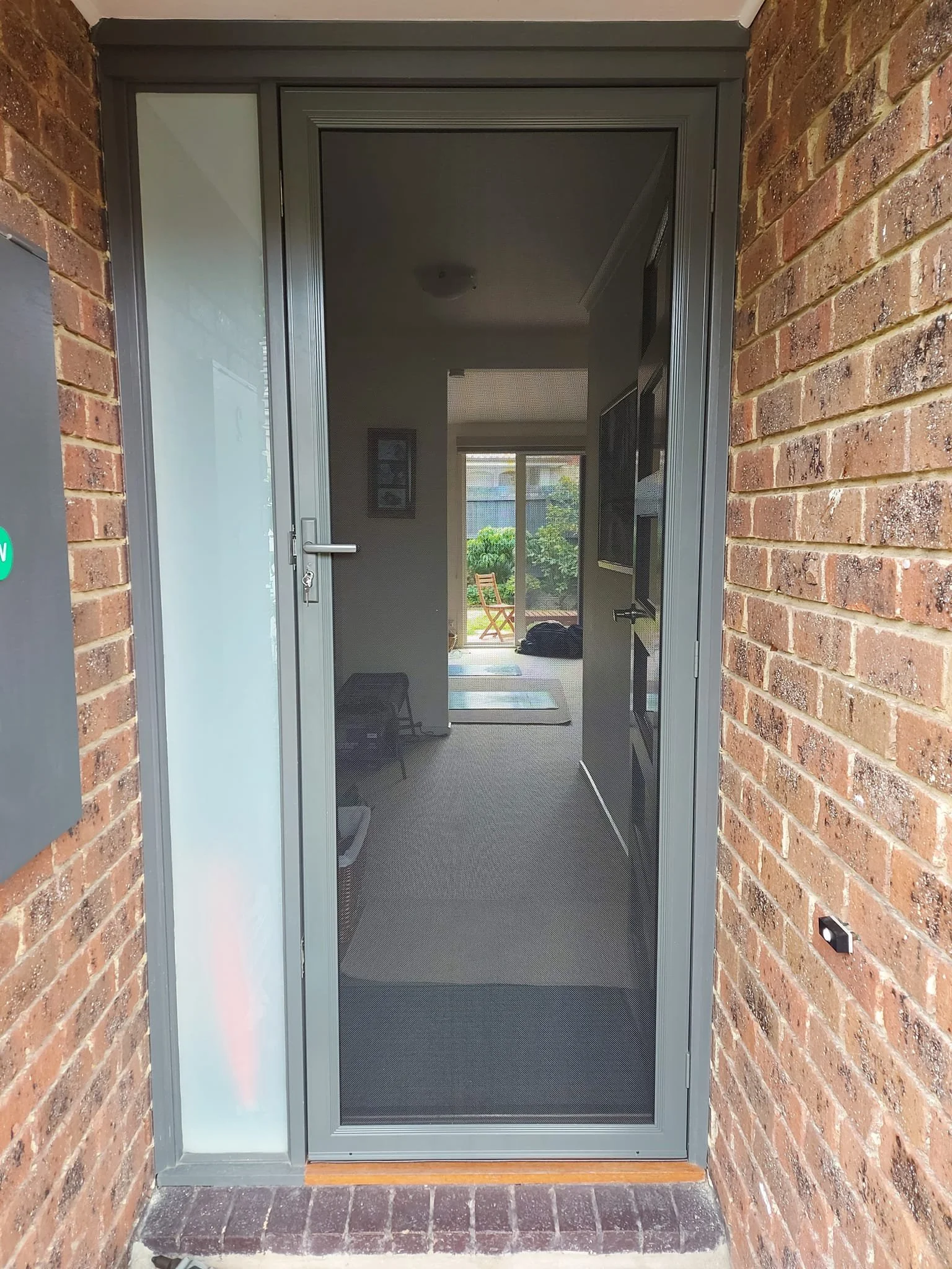 Front door with a gray screen door, brick walls, and a view of a garden with outdoor chair and plants through a sliding glass door.