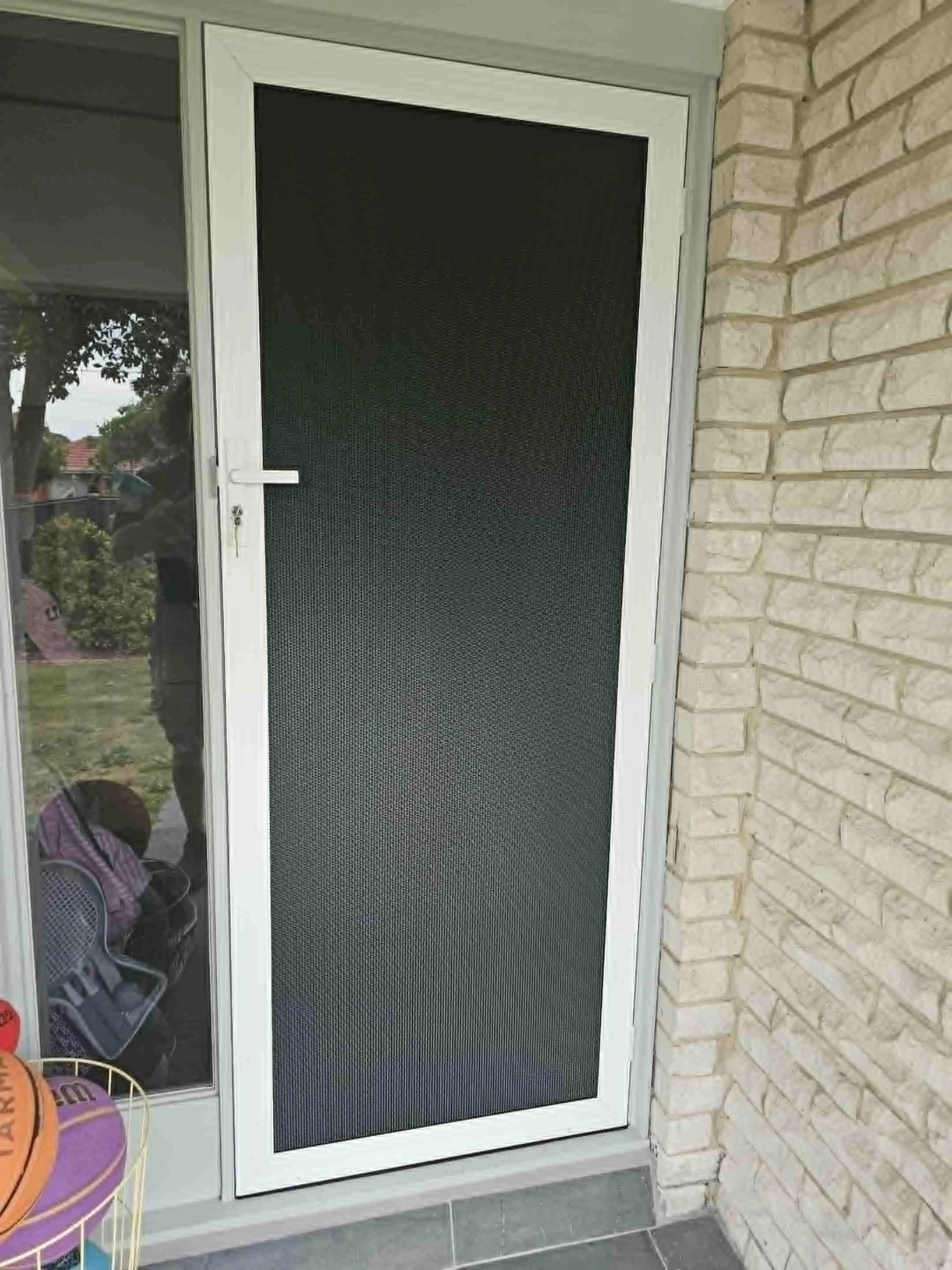 A white front door with a black screen door and a handle, next to a glass window, with a brick wall on the right side and a tiled floor.