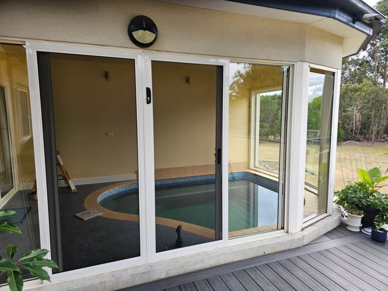 Indoor swimming pool enclosed in a screened outdoor room with sliding glass doors and large windows, located on a wooden deck with plants outside.