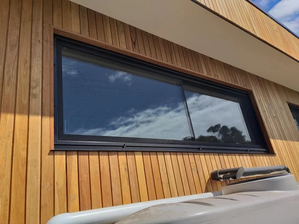 A large rectangular window on a wooden exterior wall reflecting the sky and trees.