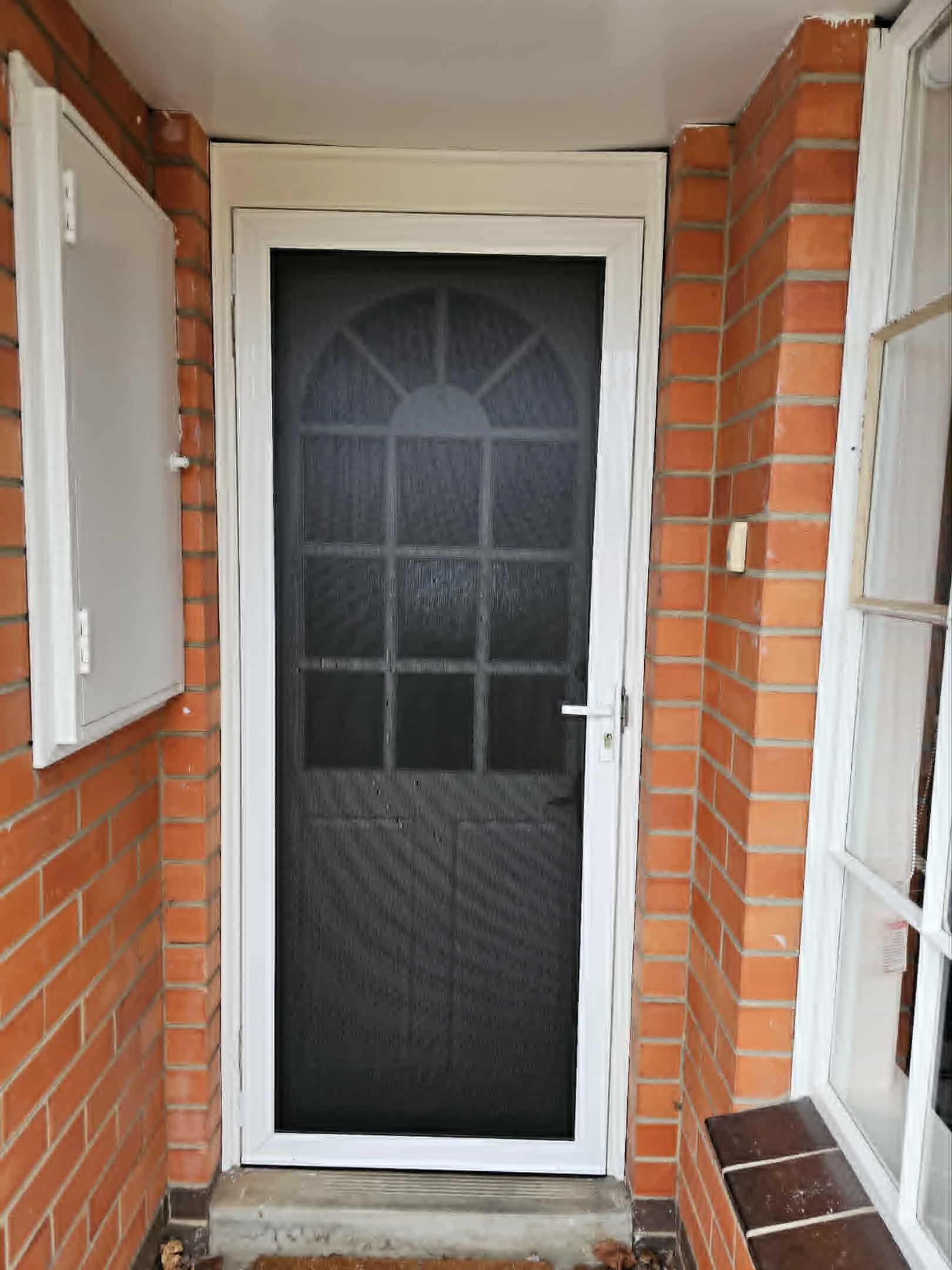 Front door with a black security screen, framed by red brick walls and white trim. To the left is an electrical box, and to the right is a window with a wide white frame. The floor has a concrete step and some fallen leaves.