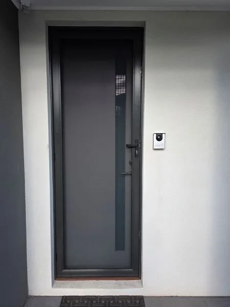 Front door made of dark glass with a black frame, on a white exterior wall, with an intercom or doorbell on the right side.