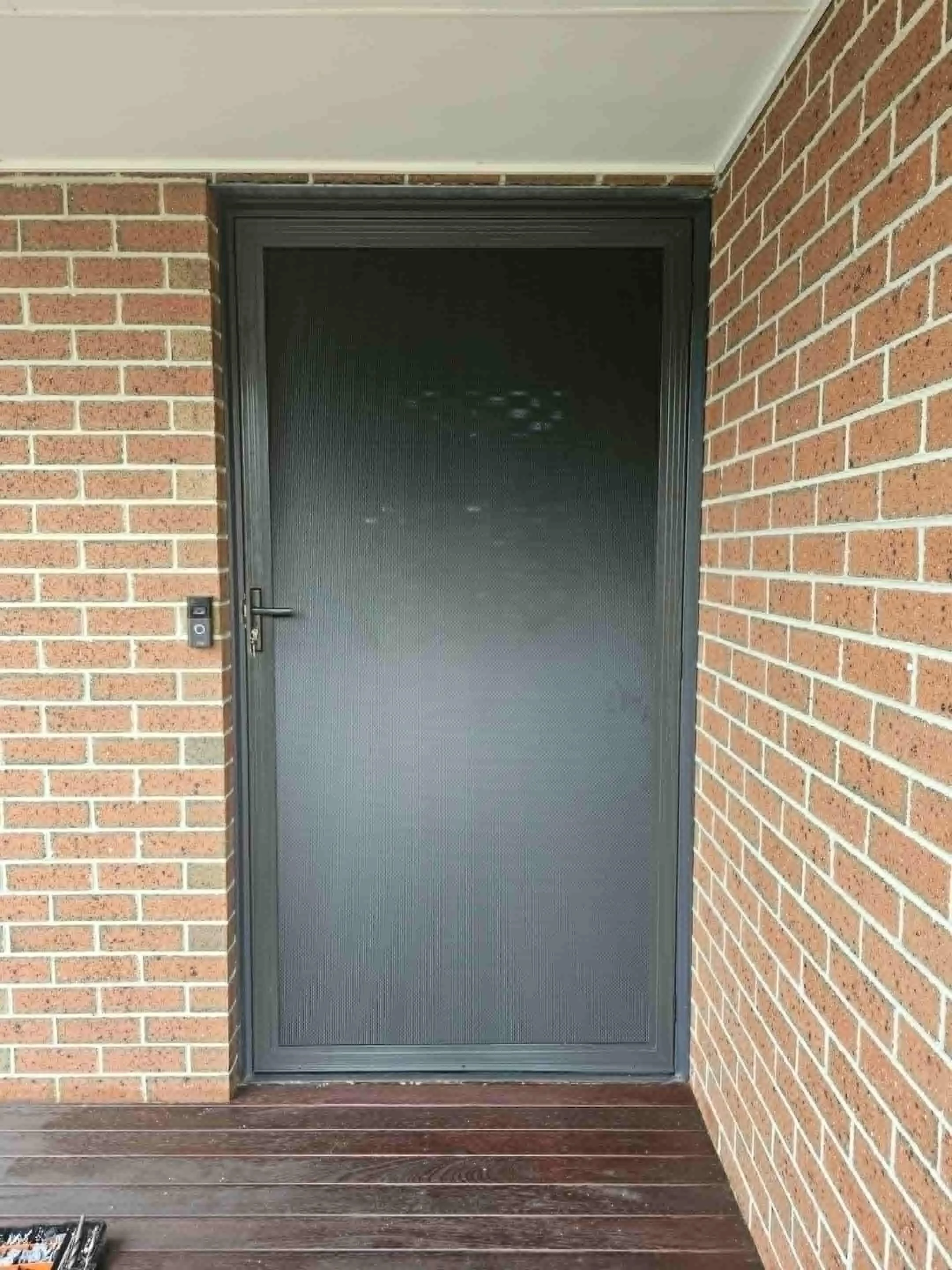 A black metal screen door on a building with brick walls and wooden porch flooring.