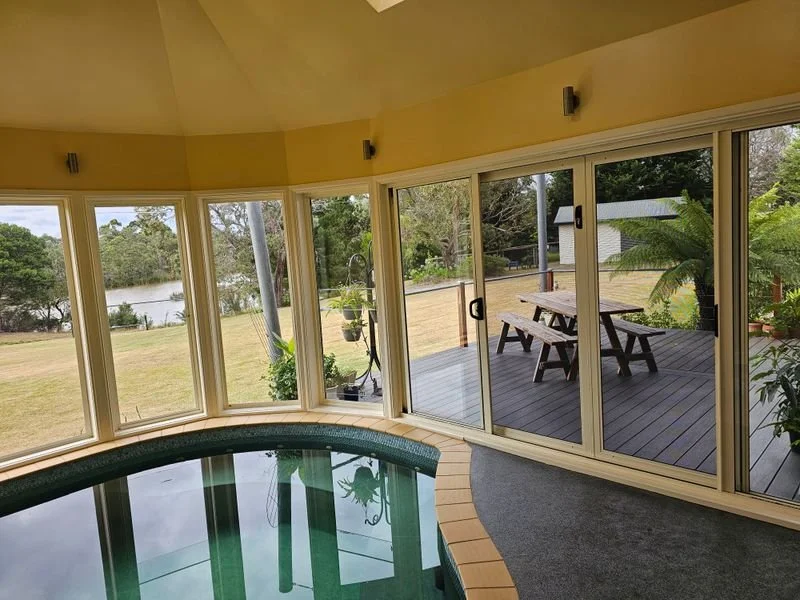 Indoor swimming pool area with large windows overlooking a grassy backyard with a pond, a wooden deck with a picnic table, and outdoor plants.