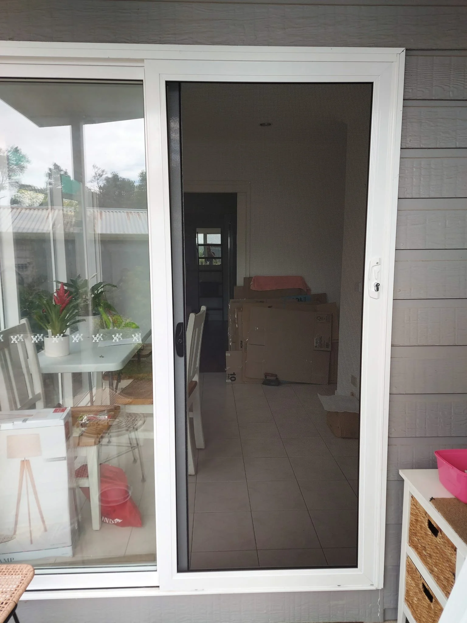 View through a sliding glass door showing an indoor dining area with a white table and chairs, and a cardboard box inside the room. The outdoor space includes a table with a plant on it and chairs, with trees and a cloudy sky in the background.