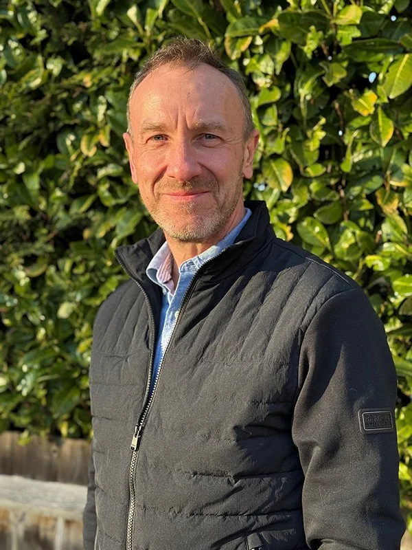 A smiling man with short hair and a beard, wearing a black quilted jacket over a collared shirt, stands outdoors in front of dense green foliage.