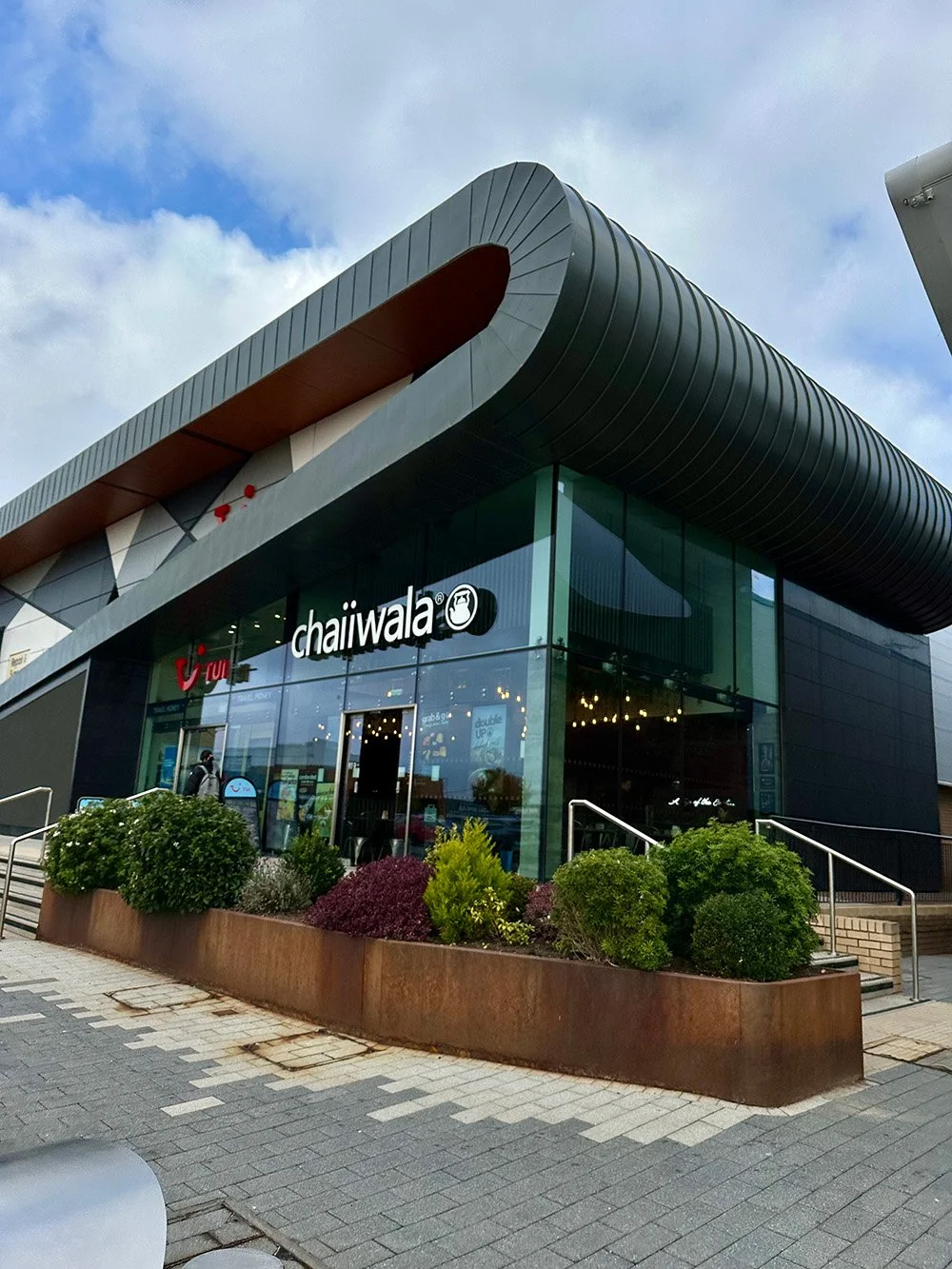 Modern commercial building with glass facade, featuring the logos of chaiwala and un limit, with landscaped greenery and a sidewalk in front.