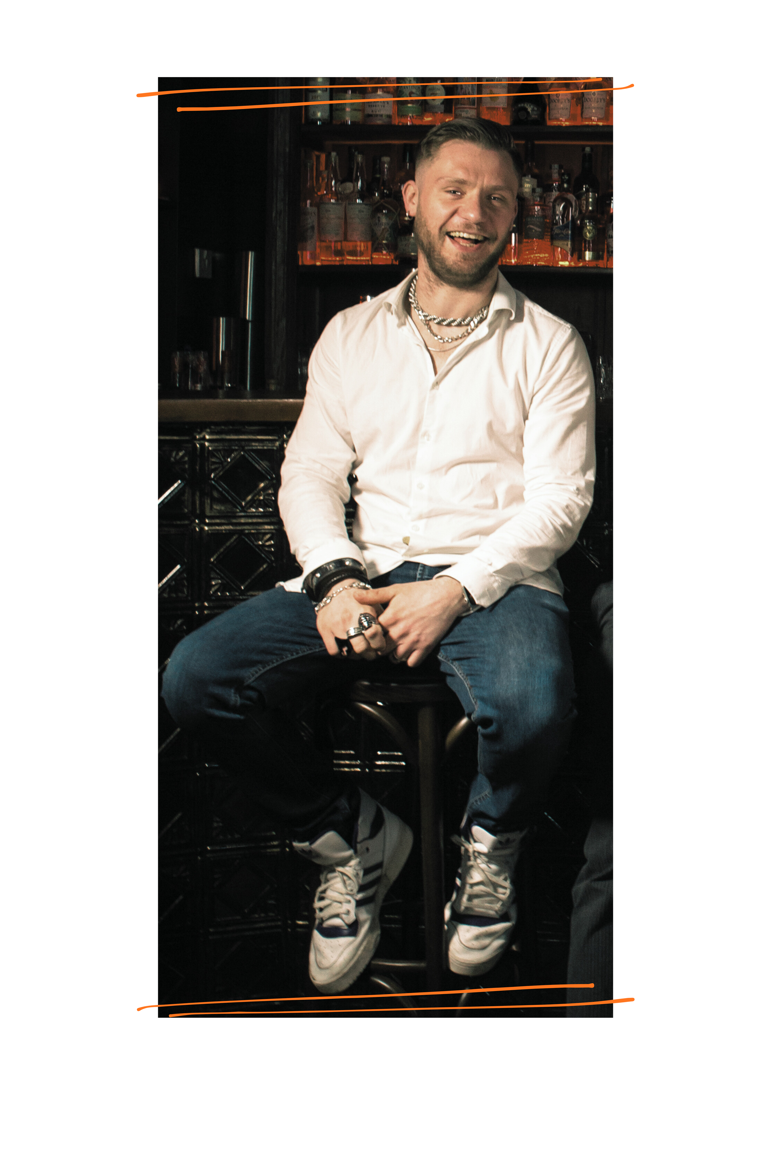 Jack, a white man with a white shirt and jeans, and chunky silver jewellery laughs at the camera