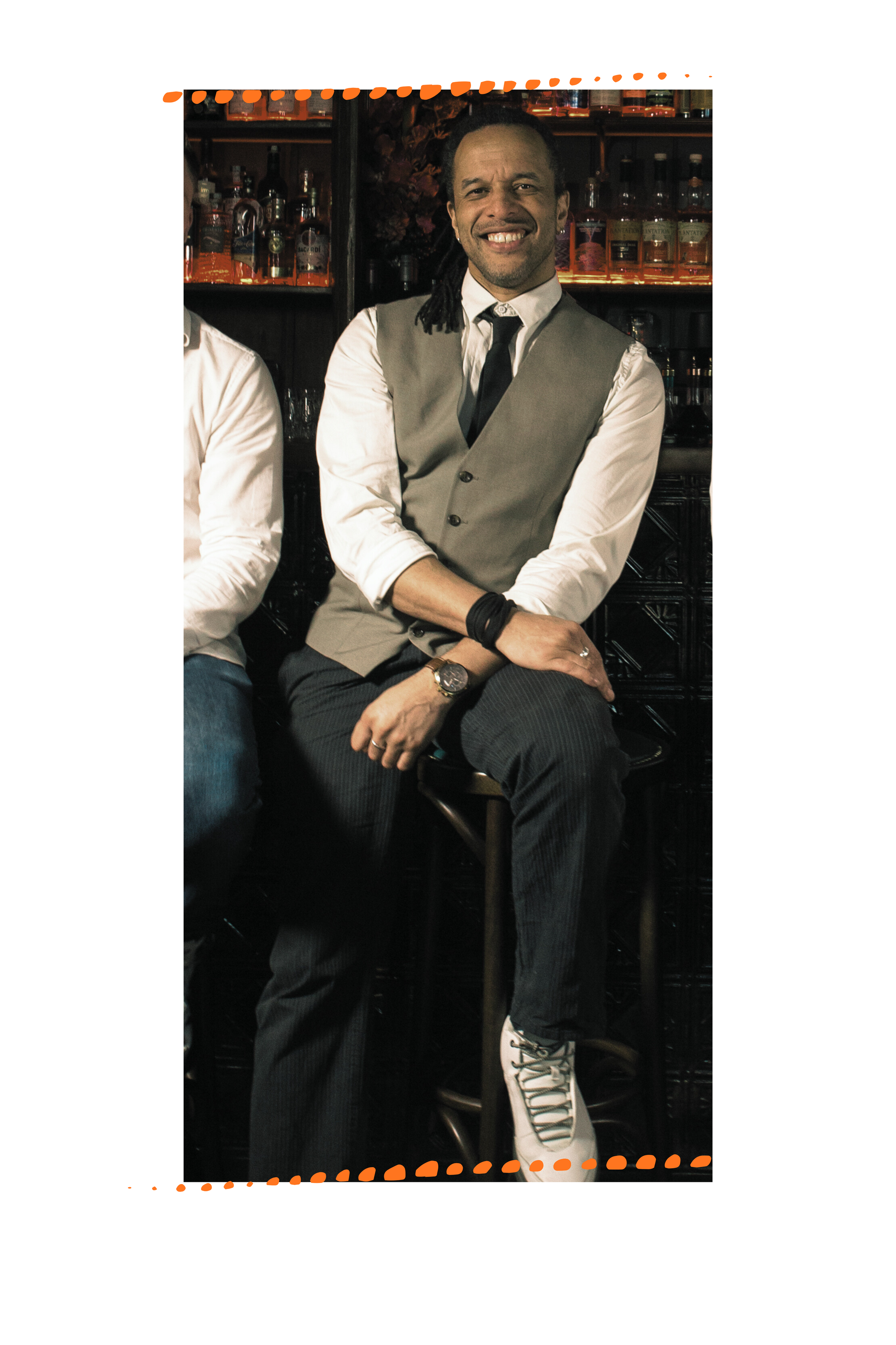 Eric, a Black man with dreads pulled up behind him sits grinning at the camera wearing a waistcoat, shirt, tie and jeans