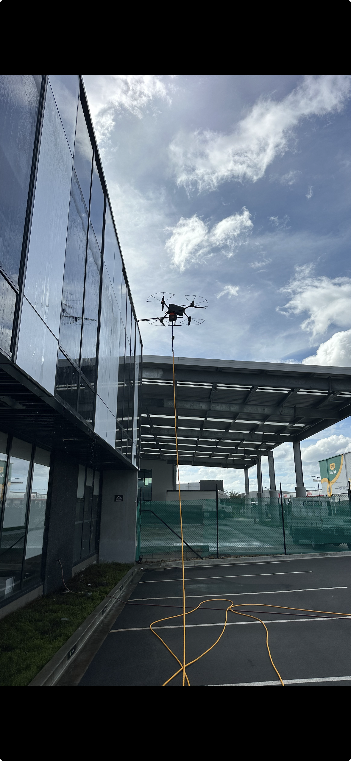 A drone is suspended in the air by a long orange cable outside a modern building with glass walls. The sky is partly cloudy with blue skies visible.