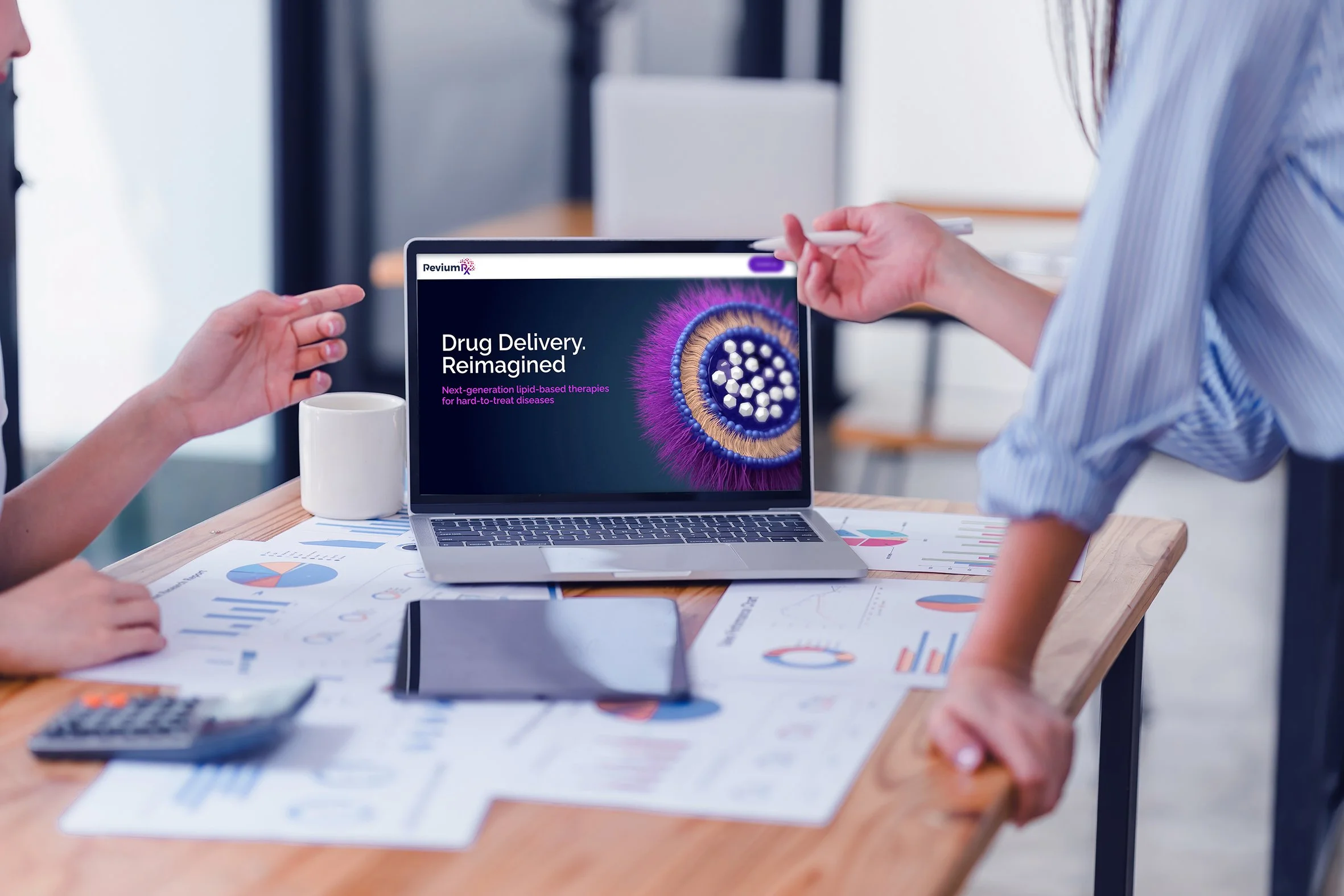 Two people discussing in a meeting room with a laptop displaying a presentation titled 'Drug Delivery. Reimagined' and an image of a microscopic view of a drug molecule; there are papers, graphs, a calculator, a tablet, and a mug on the table.