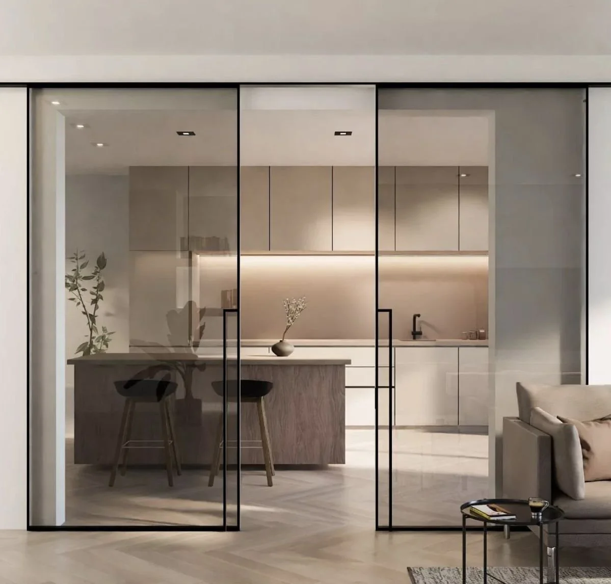 Interno di una cucina moderna con pareti di vetro trasparenti che separano la zona soggiorno. La cucina presenta mobili chiari, un'isola di legno scuro con sgabelli, e decorazioni minimaliste con un vaso di fiori. La zona soggiorno ha un divano beige e un tavolino con riviste e bicchiere.