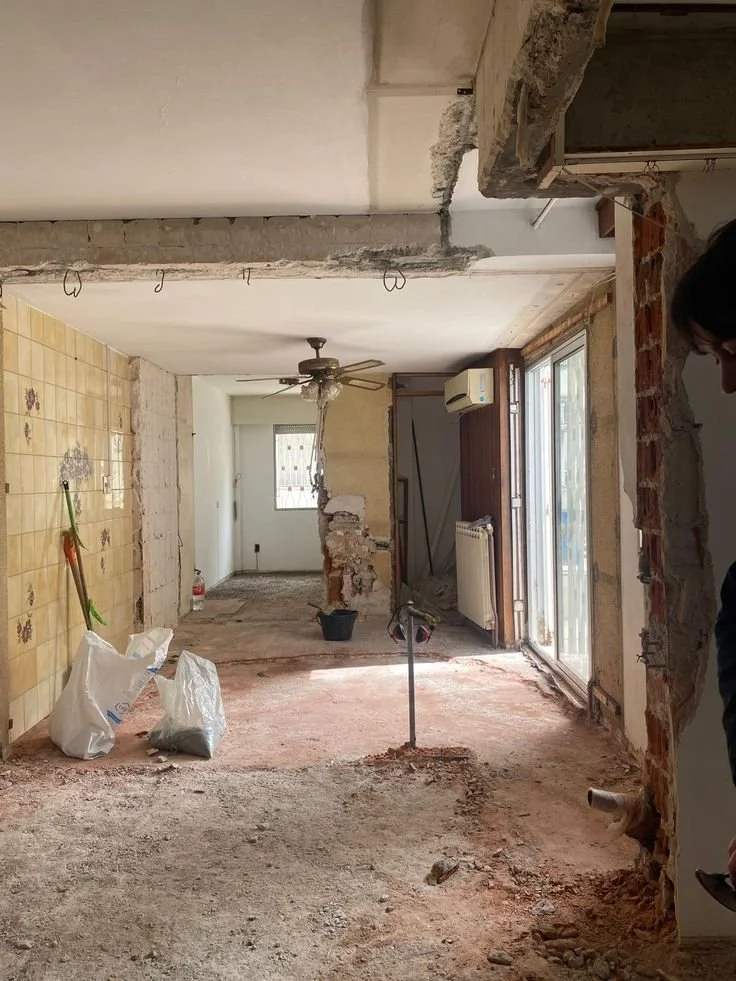 Interno di una casa in ristrutturazione con un muro parzialmente demolito, pavimento in cattive condizioni, armadio a parete, ventilatore a soffitto e grandi finestre con porta scorrevole.