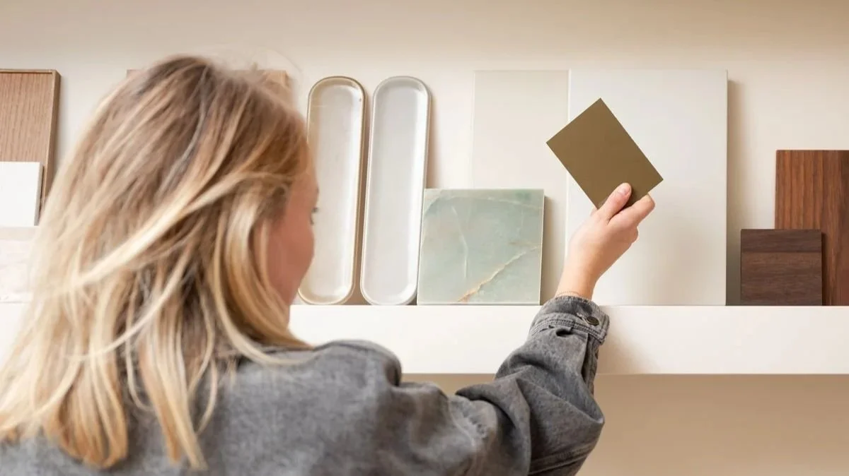 Una donna con i capelli biondi chiari che tiene un cartoncino grigio in mano, di fronte a una parete con vari campioni di materiali, tra cui ceramica, legno e marmo.