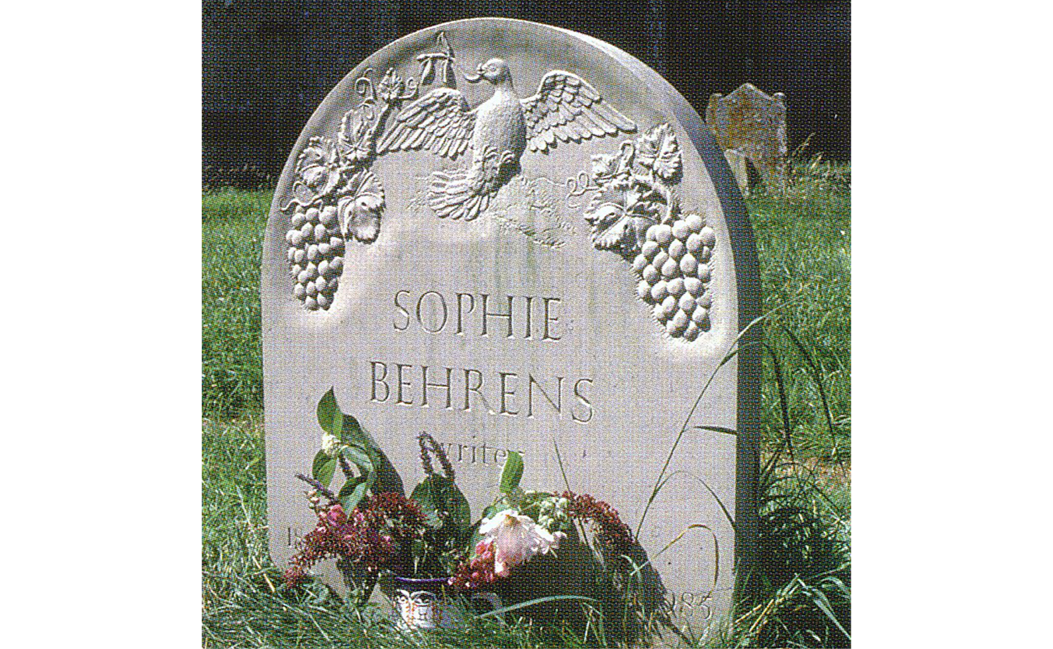 Gravestone with carved grapes and a dove, inscribed with the name 'Sophie Behrens' and the word 'writer'. Flowers and plants at the base of the gravestone.