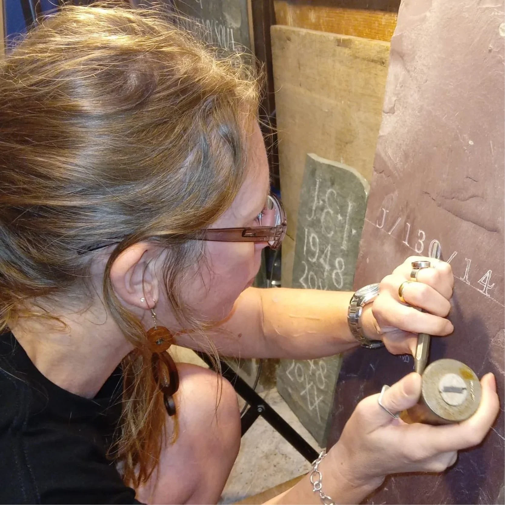 A woman with curly reddish-brown hair wearing glasses, rings, and a bracelet, appears to be engraving or working on a coin or medal with a small tool.