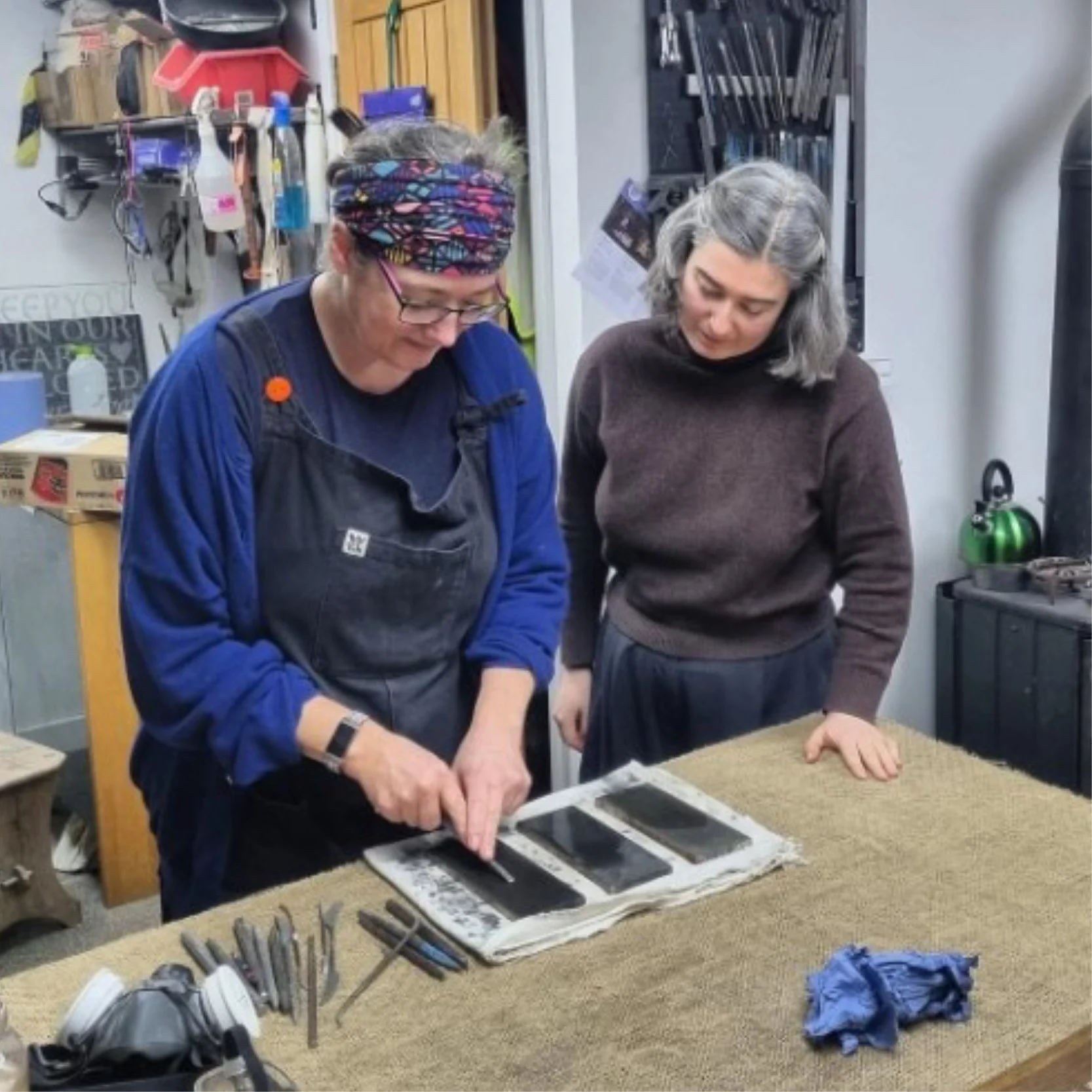 Two women working together on a craft or art project at a table in a workshop or studio, with various tools and supplies in the background.