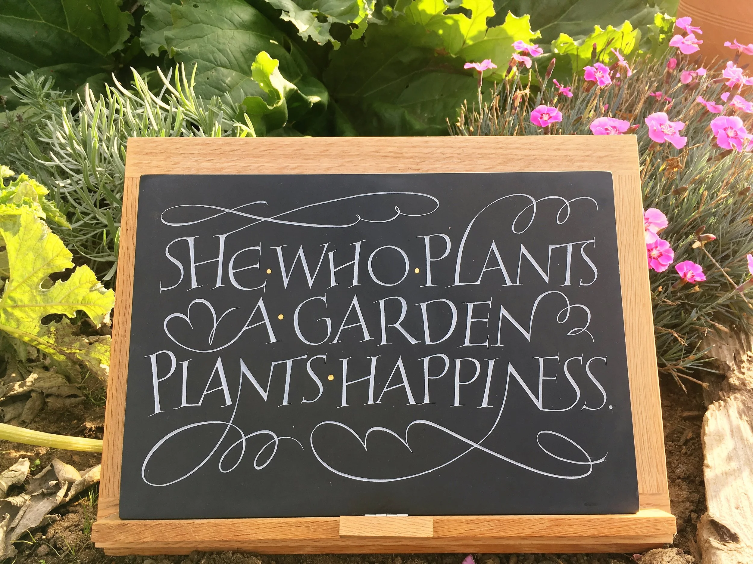 A black chalkboard sign with white cursive writing that mentions she, who plants, and happiness, surrounded by pink and purple flowers and green foliage in a garden.