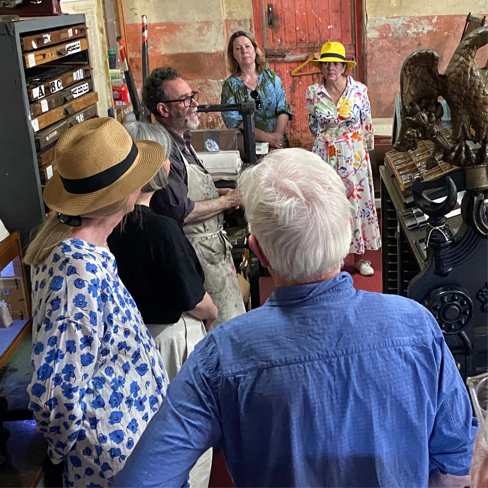 A group of people gathered in an art studio or gallery, listening to a presentation or demonstration. The setting has an industrial, rustic style with worn walls, wooden doors, and sculptures on display.