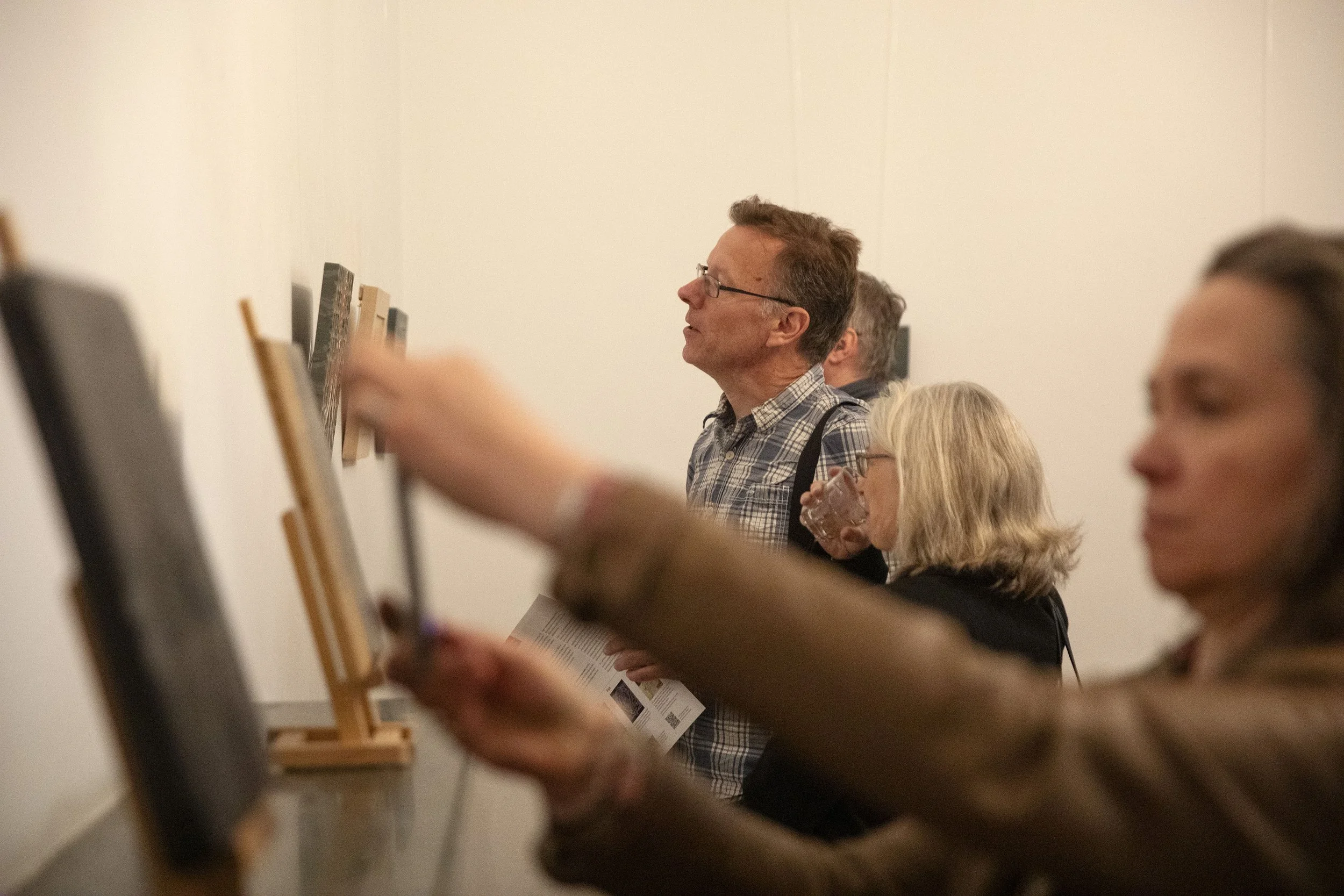 People observing artwork or exhibits in a gallery or museum, with a man in a plaid shirt and glasses in the center.