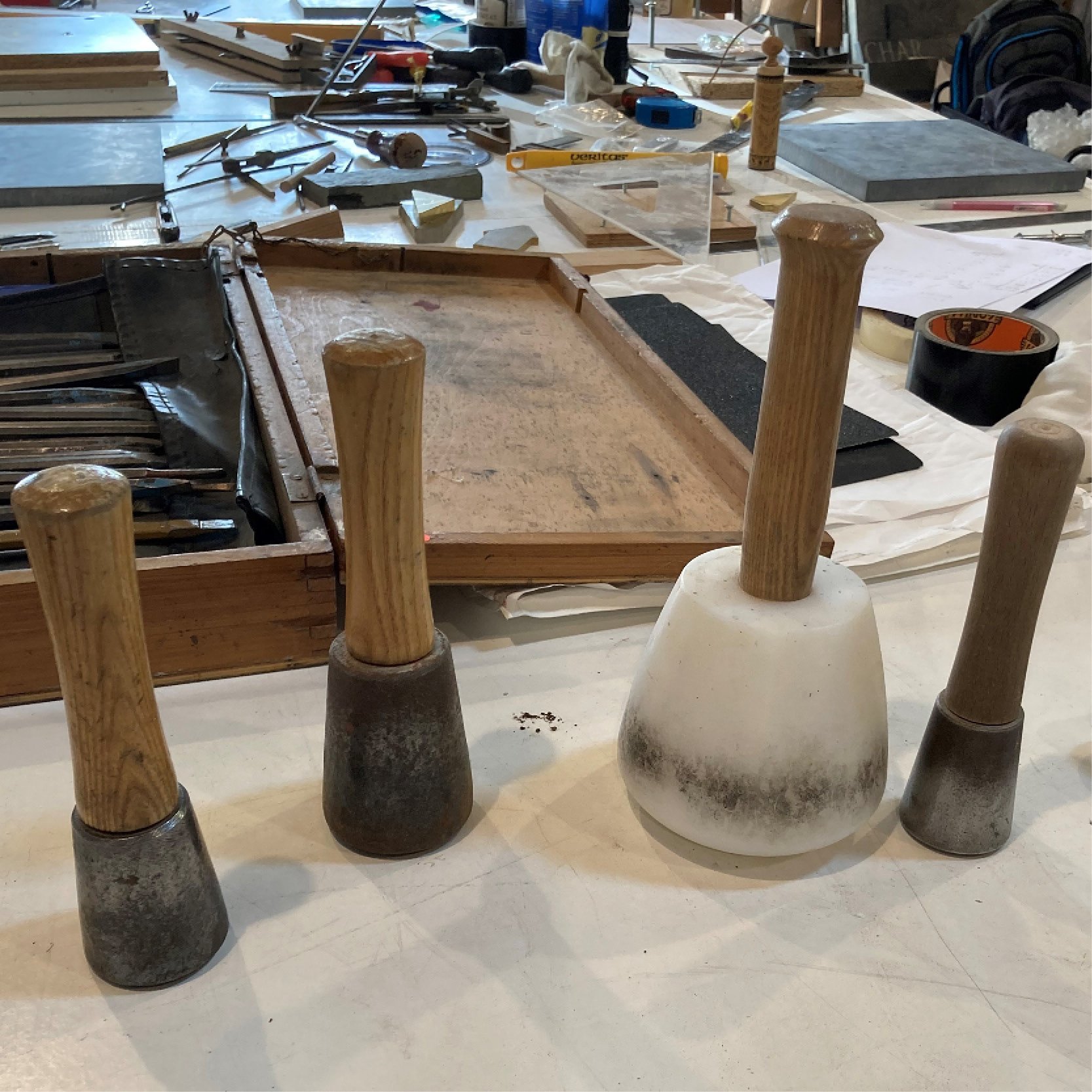 A workshop or studio space featuring a table with various tools, materials, and sketches for woodworking or furniture making. Four chairs are turned upside down on the foreground, showing their wooden legs and bases.