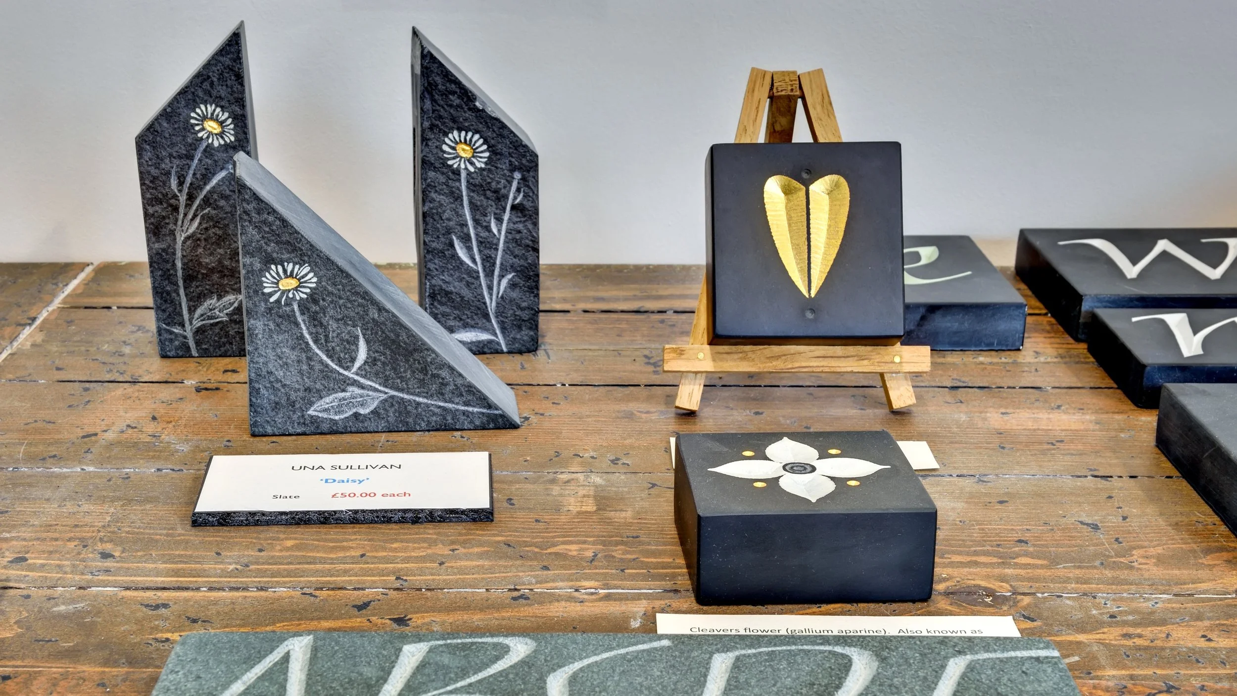 Various painted and engraved stones, including black stones with daisy flowers and white flower designs, displayed on a wooden table with a small wooden easel and a name tag that reads 'Daisy' priced at 50 pounds each.