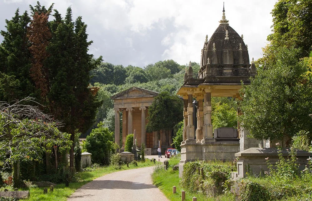 A peaceful cemetery pathway lined with trees and historical tombs, featuring a domed mausoleum and classical-style buildings in the background.