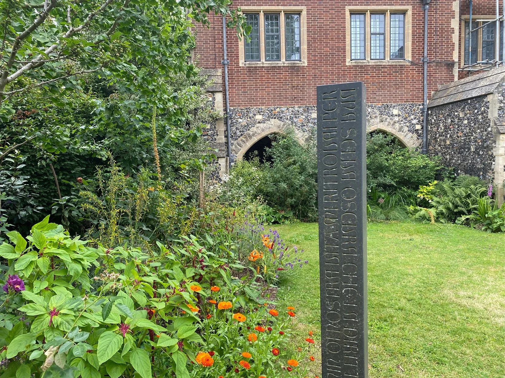 A garden with a variety of green plants and colorful flowers in front of a brick building. A dark gray stone pillar with engraved text is in the foreground.