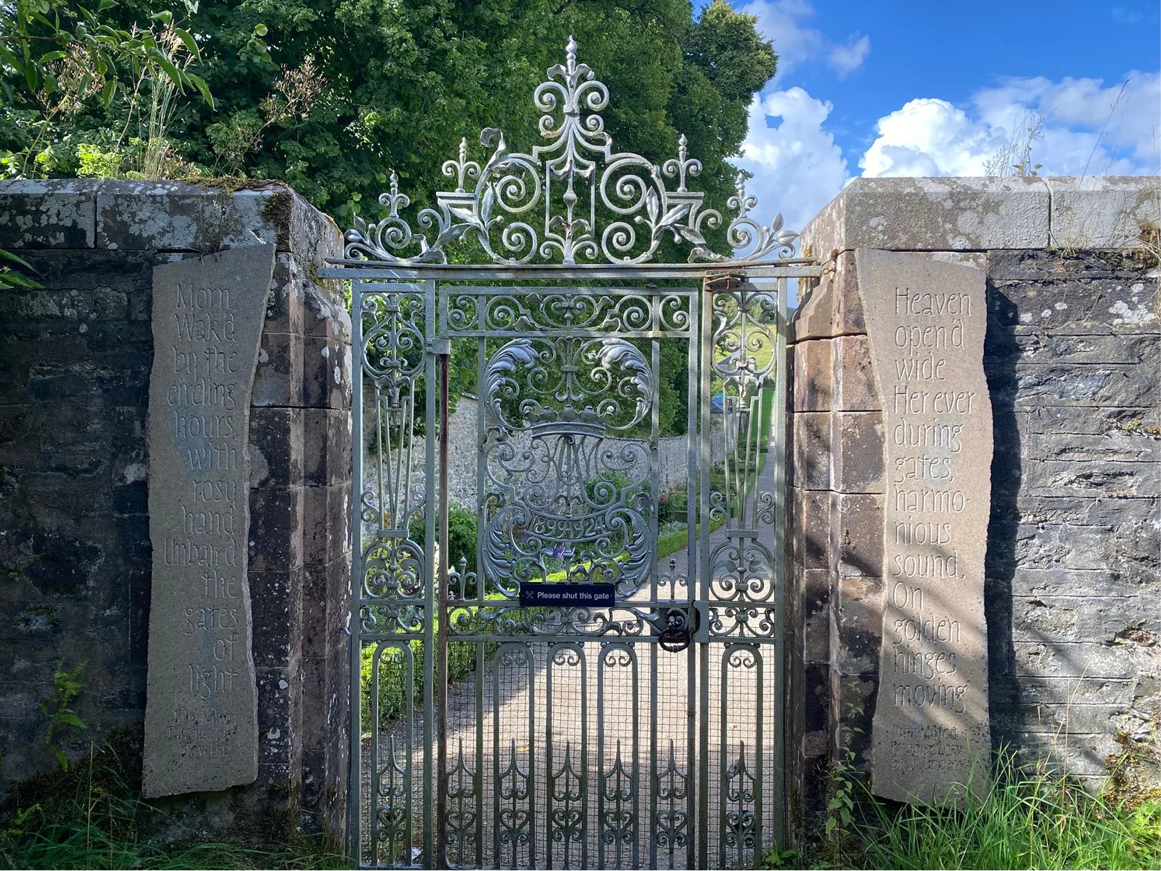 An ornate iron gate flanked by stone pillars with engraved poetry, opening to a garden with trees and a blue sky with white clouds.