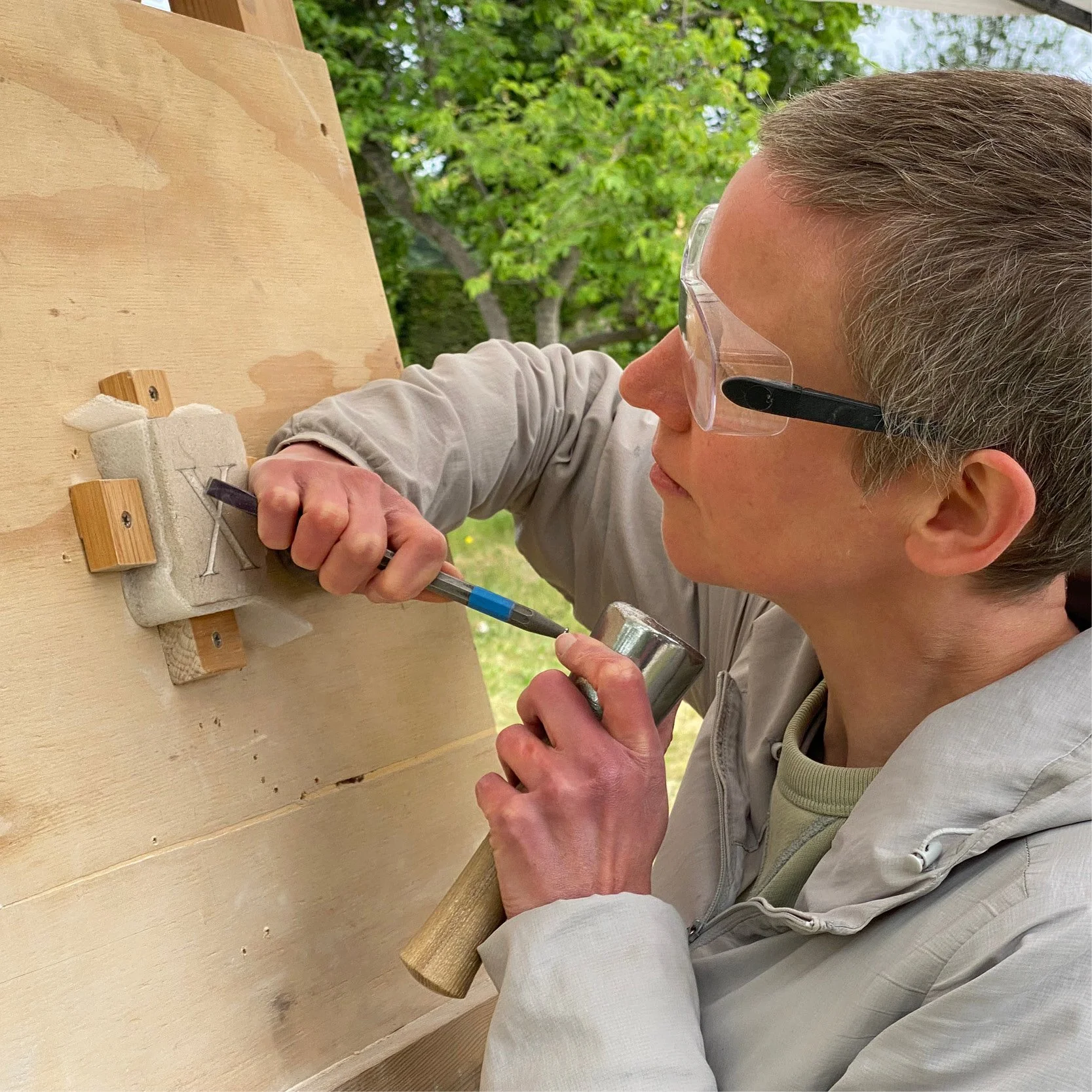 A person wearing safety glasses using a hammer and chisel to carve a letter 'V' into a stone block outside on a wooden workbench.