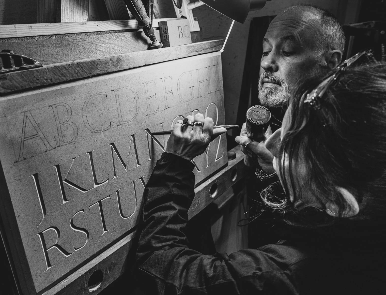 Two people working on laser engraving a wooden board with the alphabet in uppercase letters, in a workshop. One person is closely observing, while the other is holding a tool and a small container.