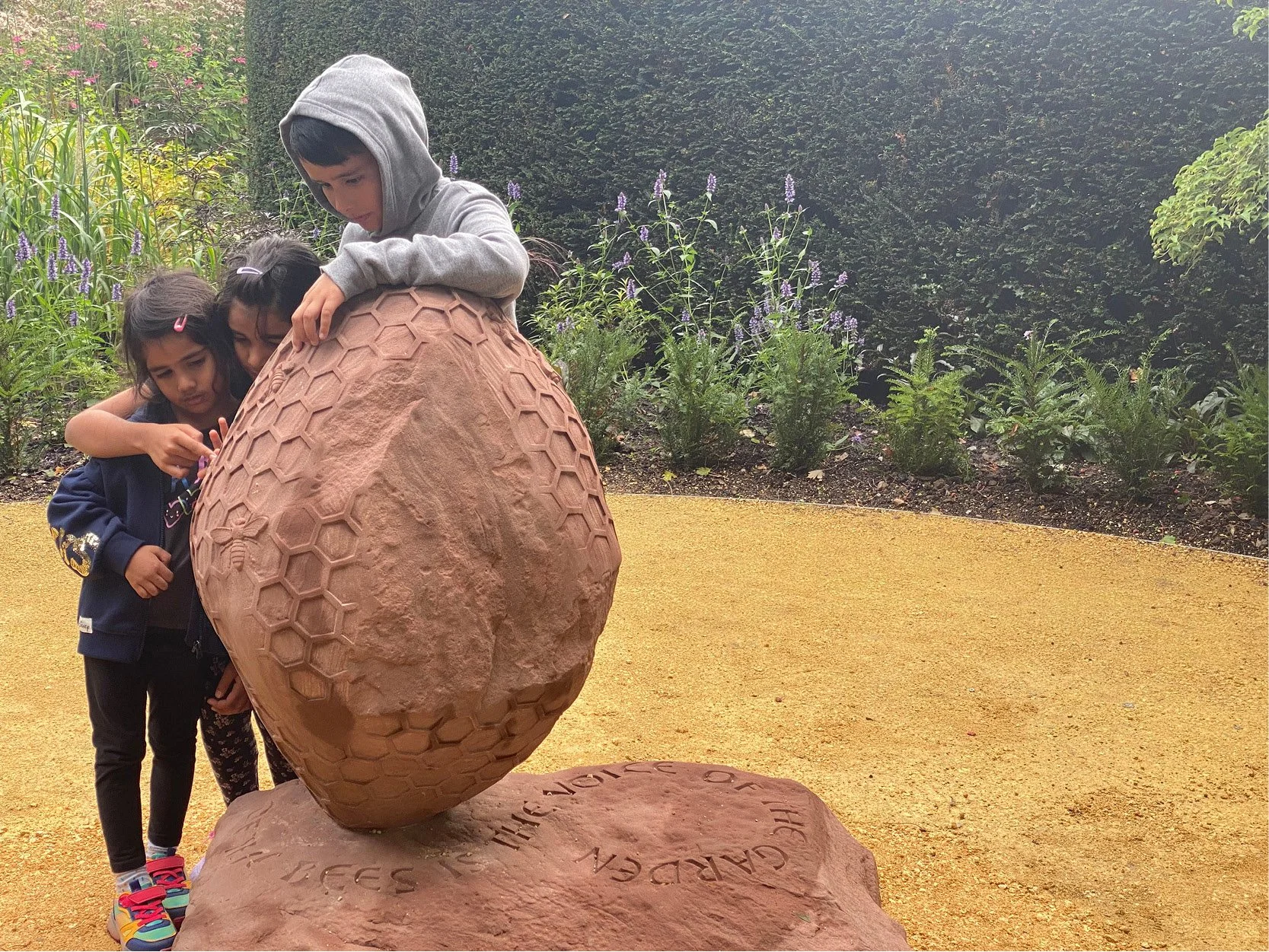 Three children observe a sculpture of a large egg-shaped object with a honeycomb pattern, set outdoors on a yellow ground with green plants and purple flowers in the background.